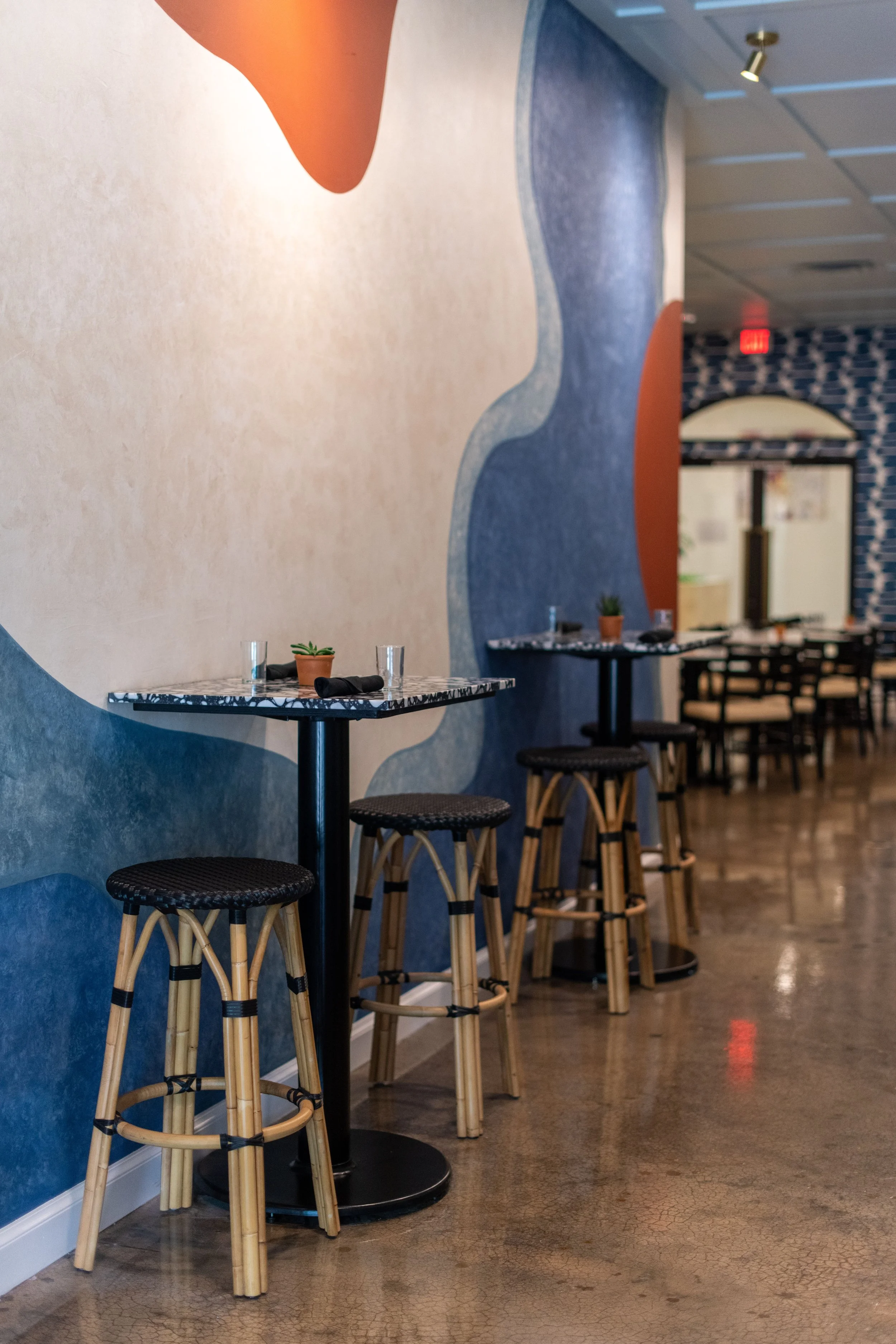 Interior of a restaurant with a line of tables and stools, blue and orange wall decor, small potted plants on tables.