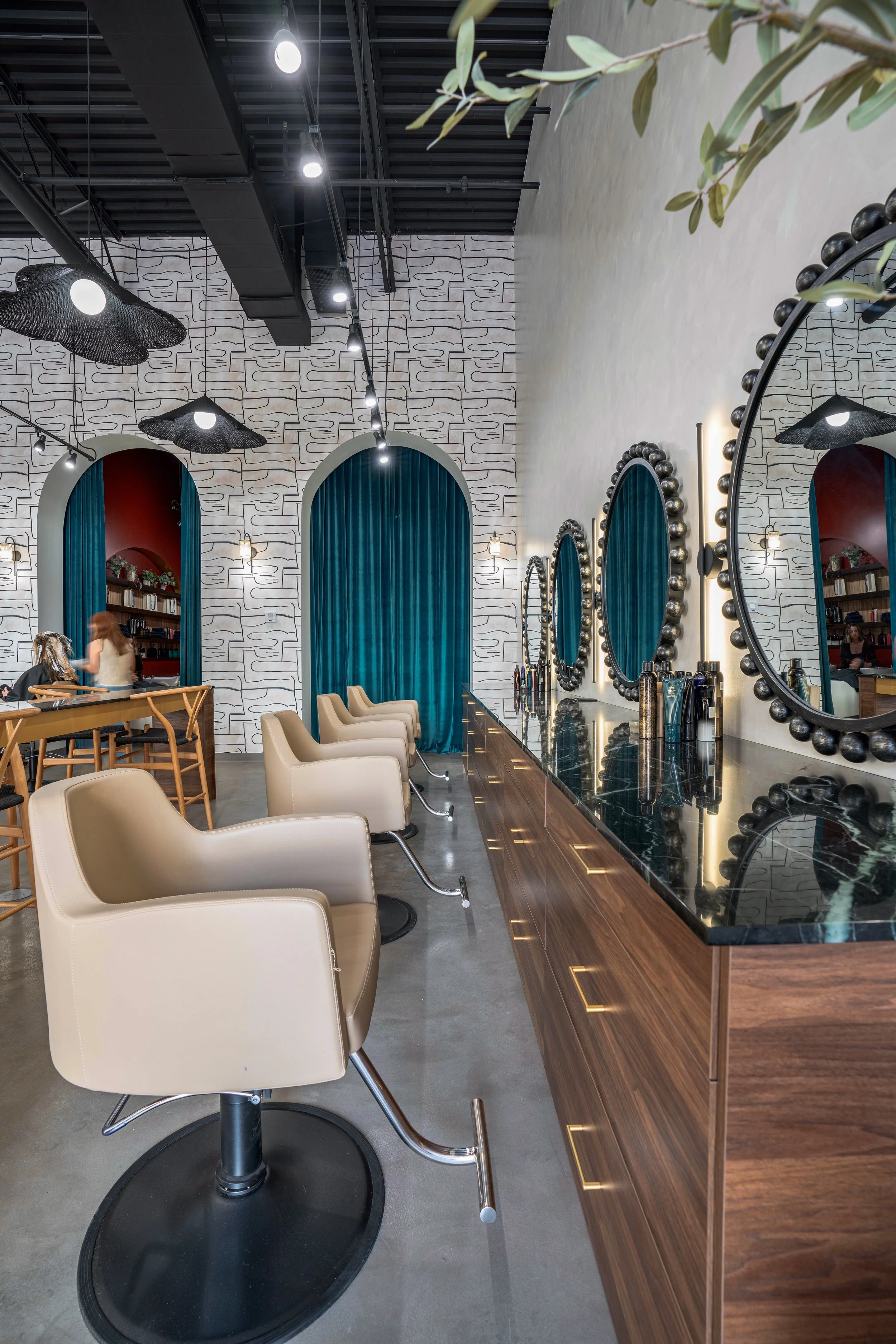 Interior of a modern hair salon with a row of beige styling chairs in front of a black marble countertop. Large round mirrors with beaded frames hang along the wall. The wall has a white patterned texture, and there are blue curtains framing arched a