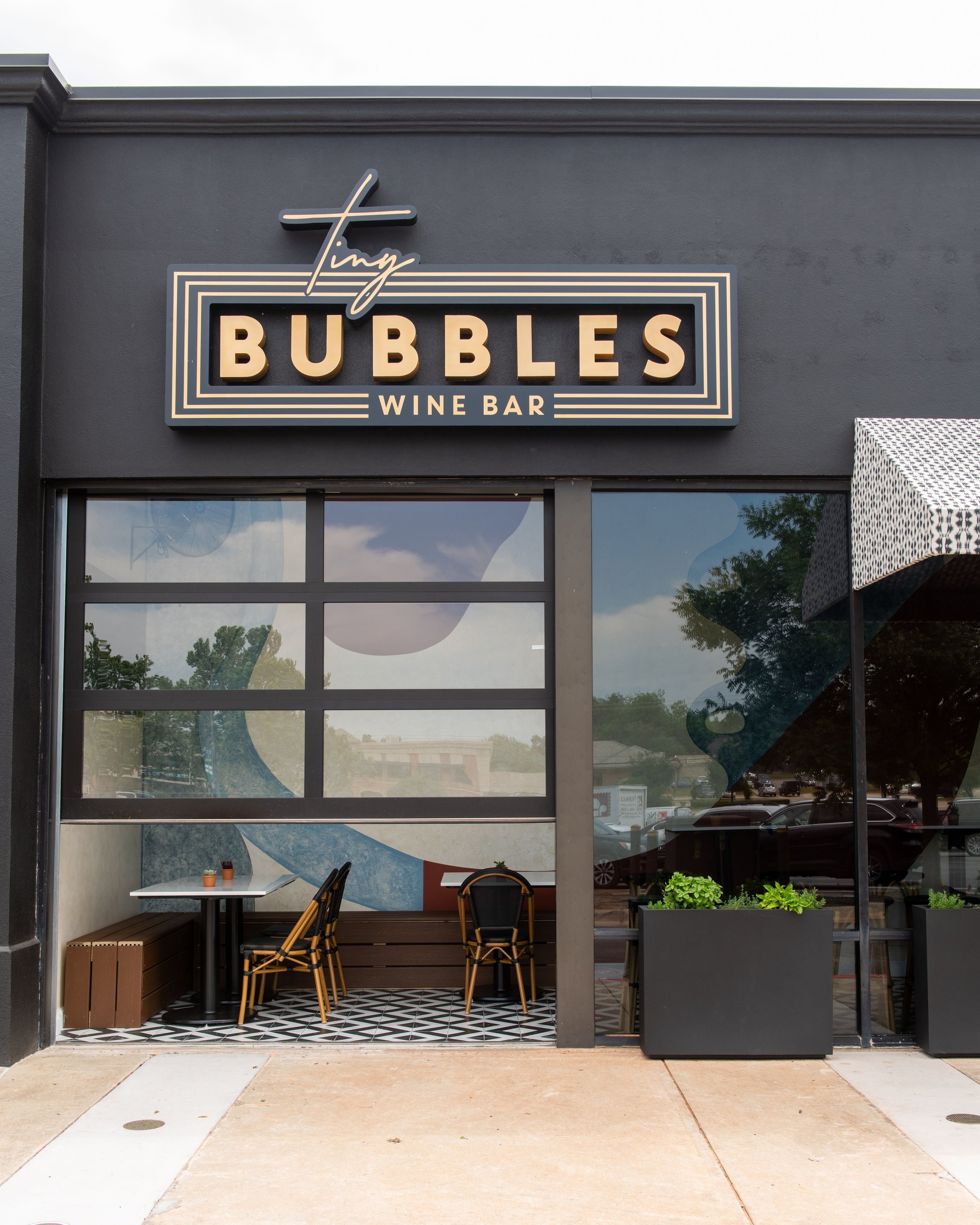 The exterior of Tiny Bubbles Wine Bar with a black signboard displaying the name in large letters, part of the entrance and seating area visible through the glass window, and outdoor decor including black planters with green plants.