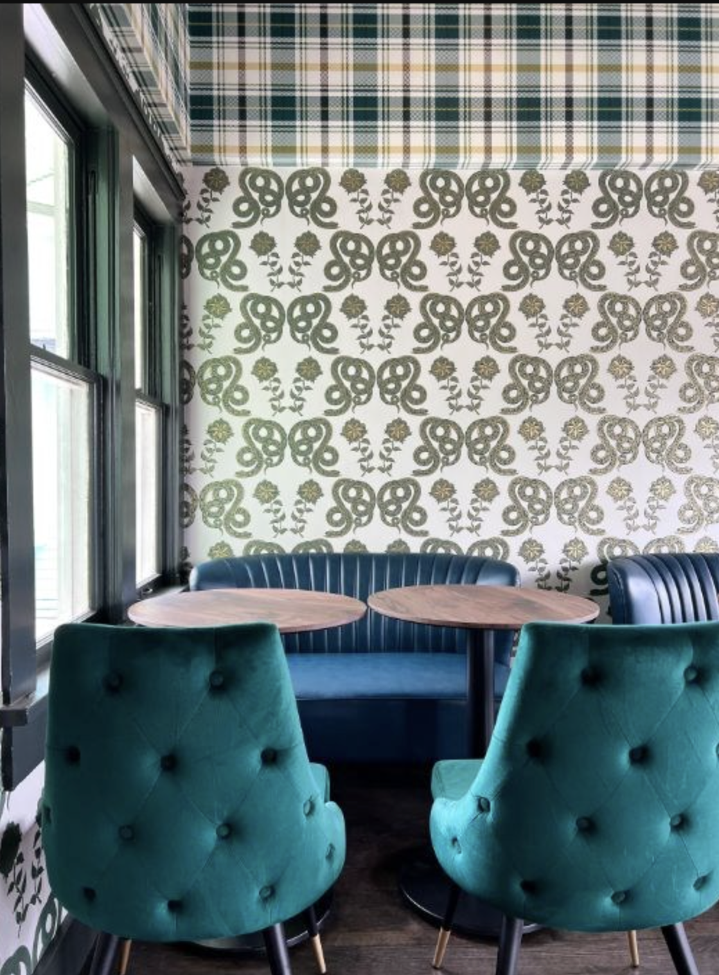 Corner of a cozy restaurant with green velvet chairs, a blue booth, wooden tables, floral wallpaper, plaid ceiling, and windows letting in natural light.