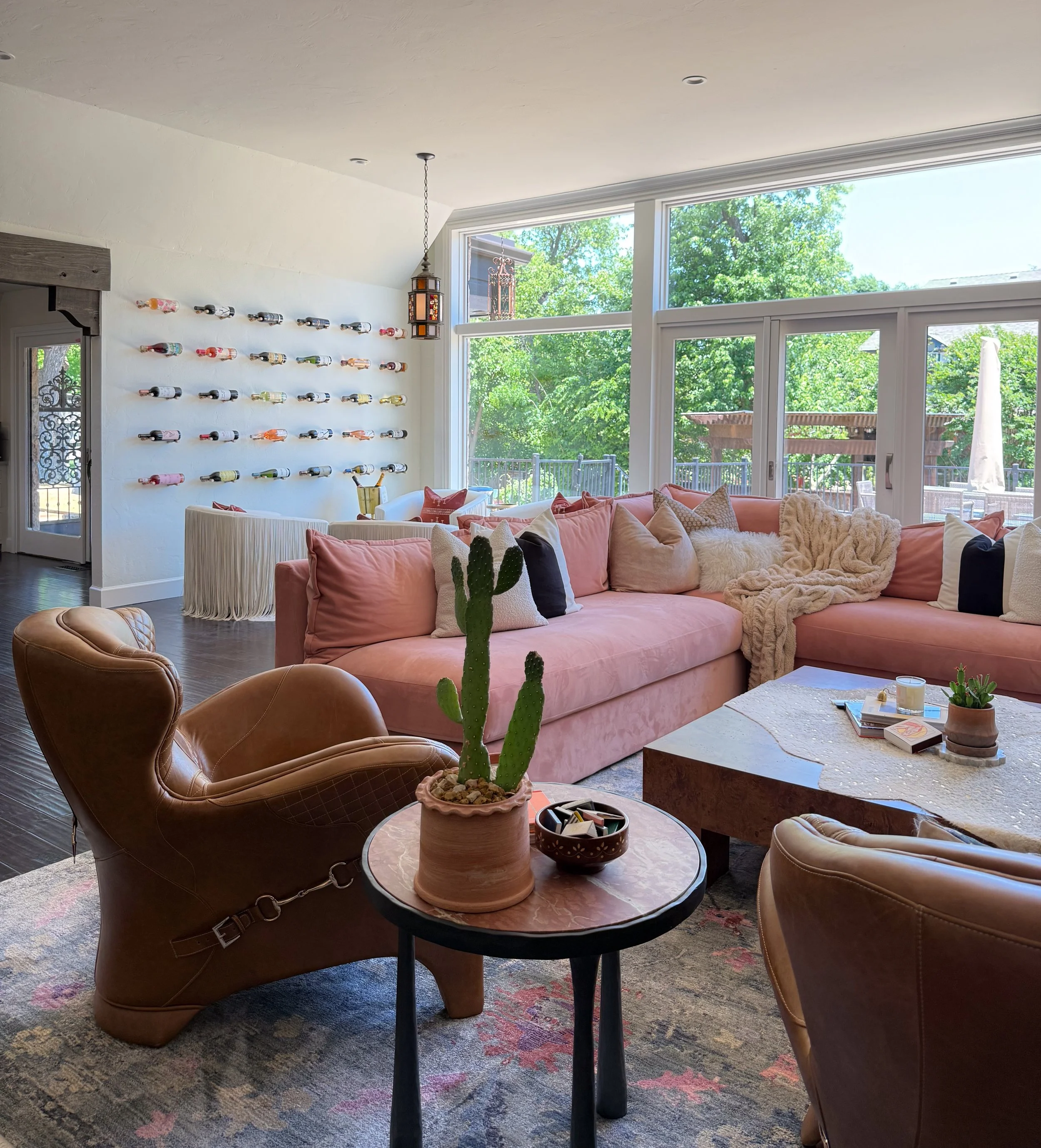 Bright living room with large windows, pink sofa with pillows, leather armchairs, wooden coffee table with books and candles, cactus plant, wall decorated with colorful wine bottles, and outdoor patio visible through windows.