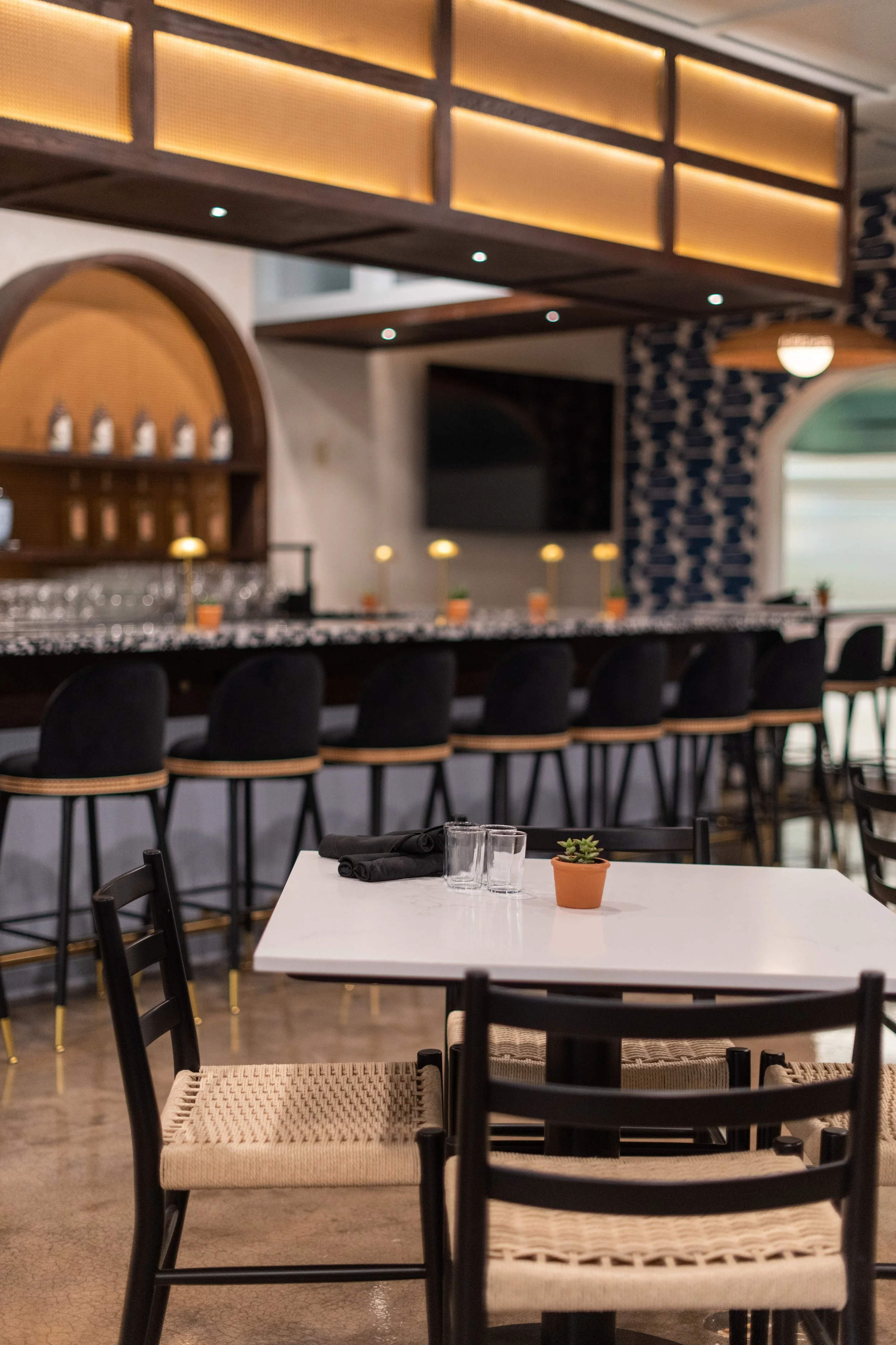 Empty restaurant with a white table, black chairs with woven seats, and a small potted succulent. The background shows a bar area with high black chairs, a marble countertop, and decorative lighting, along with a patterned wall and a television.