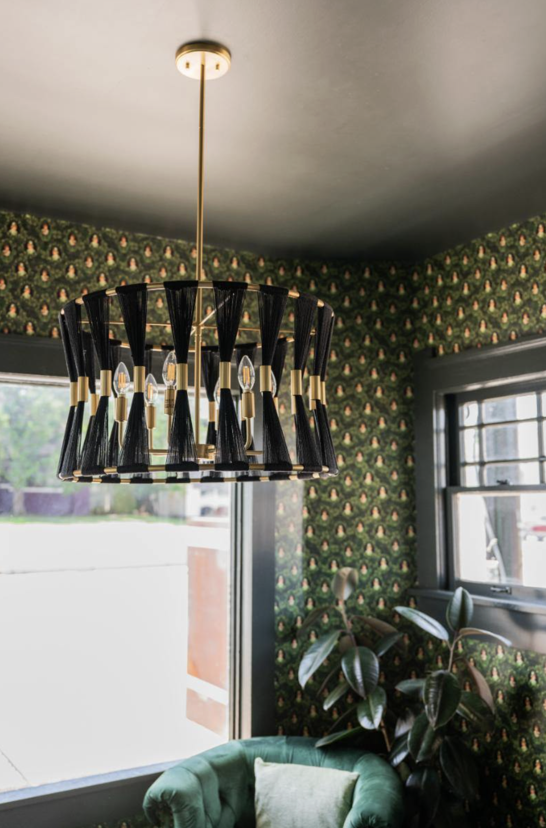 A modern dining or living room with a black chandelier featuring multiple candle-shaped light bulbs with gold accents, hanging from a gold ceiling fixture. The room has green floral wallpaper, a large window with black trim, a green armchair, and a p