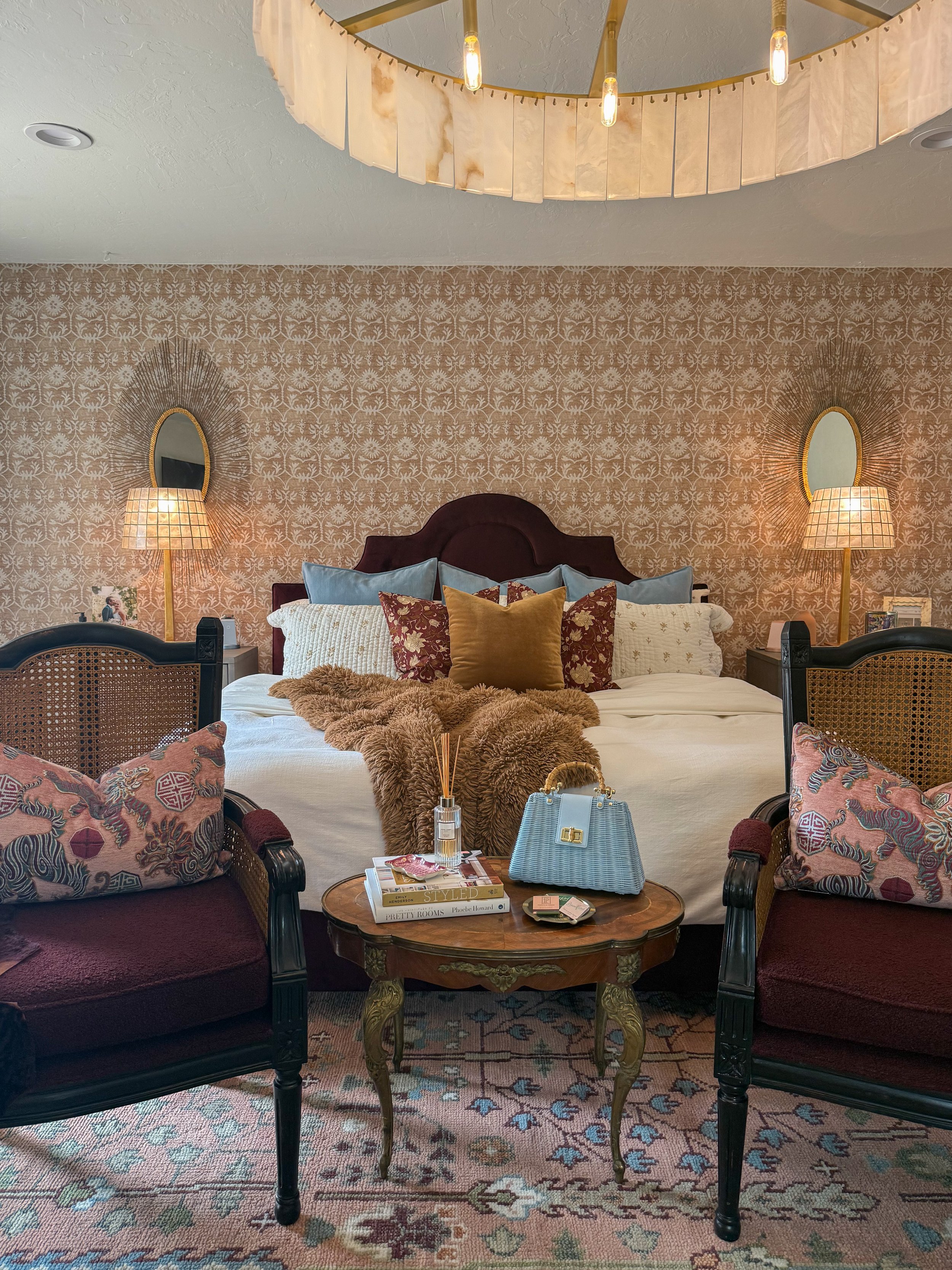 A cozy bedroom with a large bed, decorated with multiple pillows and a fur throw. There are two chairs with patterned cushions, a wooden coffee table holding magazines, a handbag, and decorative items. The wallpaper has a delicate floral pattern, and