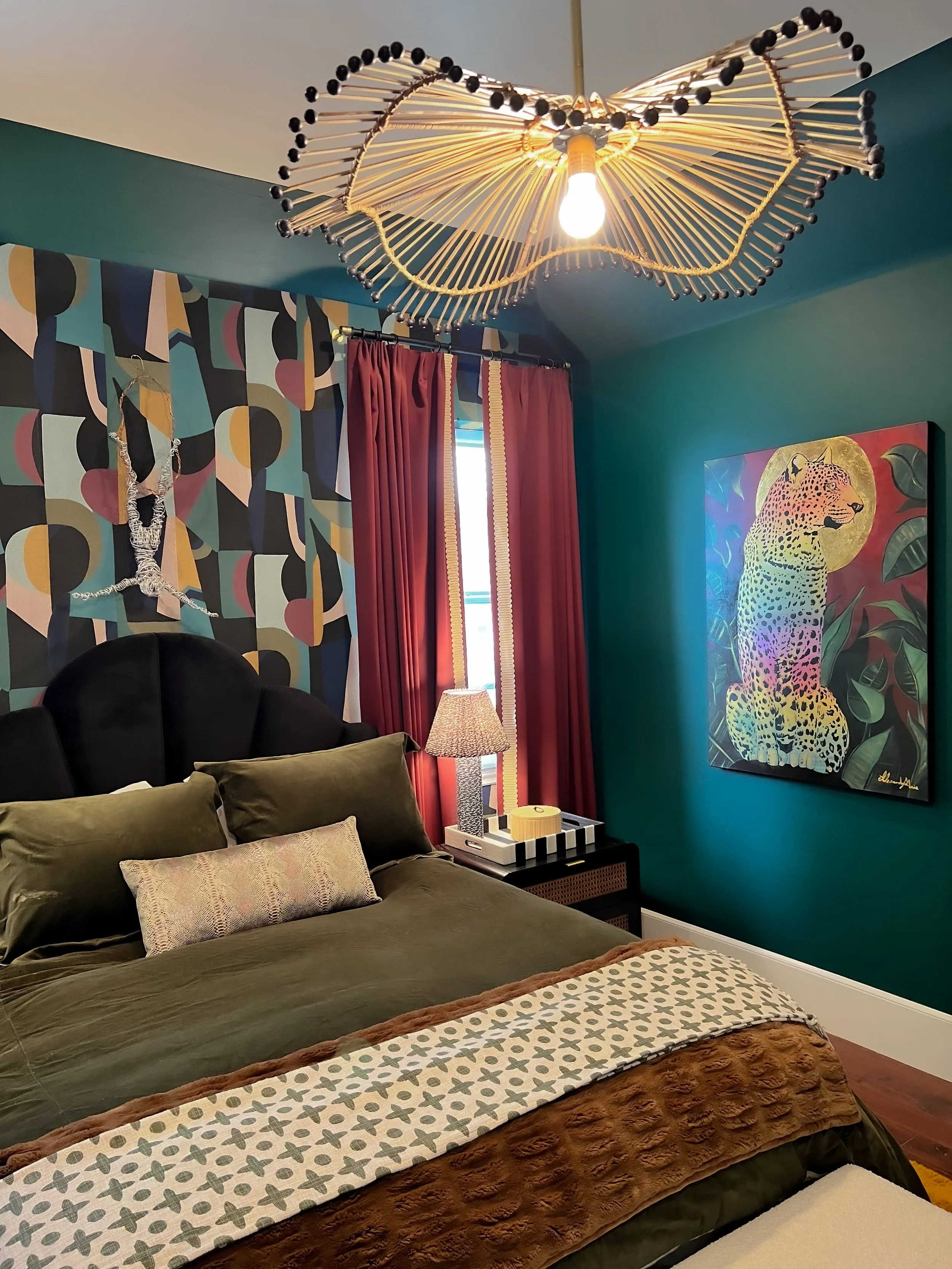 A bedroom with dark green walls, a bed with olive and patterned pillows, and a headboard. There is a colorful, geometric wallpaper behind the bed, a window with red curtains, a bedside table with a lamp, and a vibrant painting of a leopard on the wal