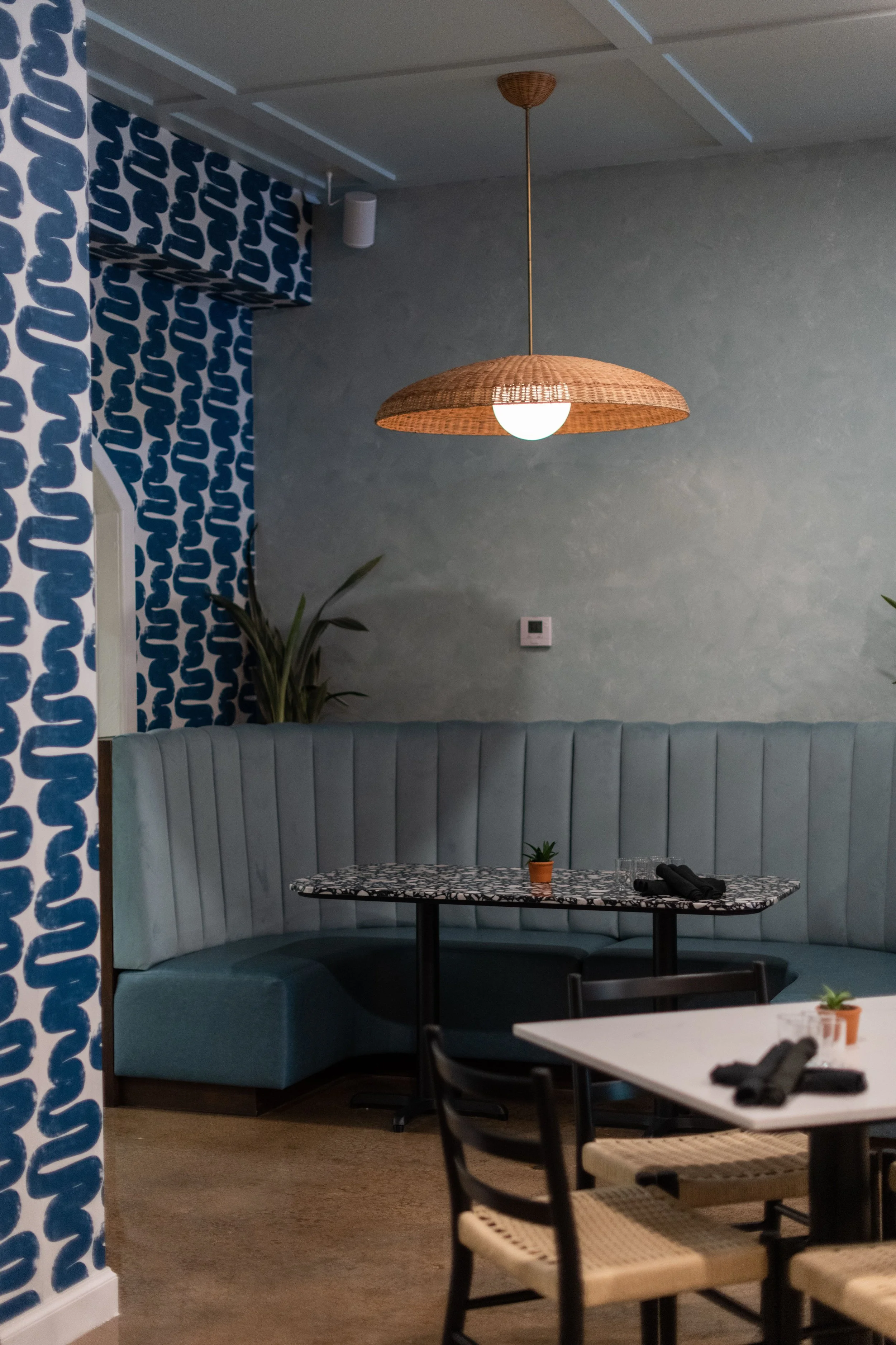 Interior view of a modern restaurant with a curved booth, patterned tables, potted plants, and a rattan pendant light.