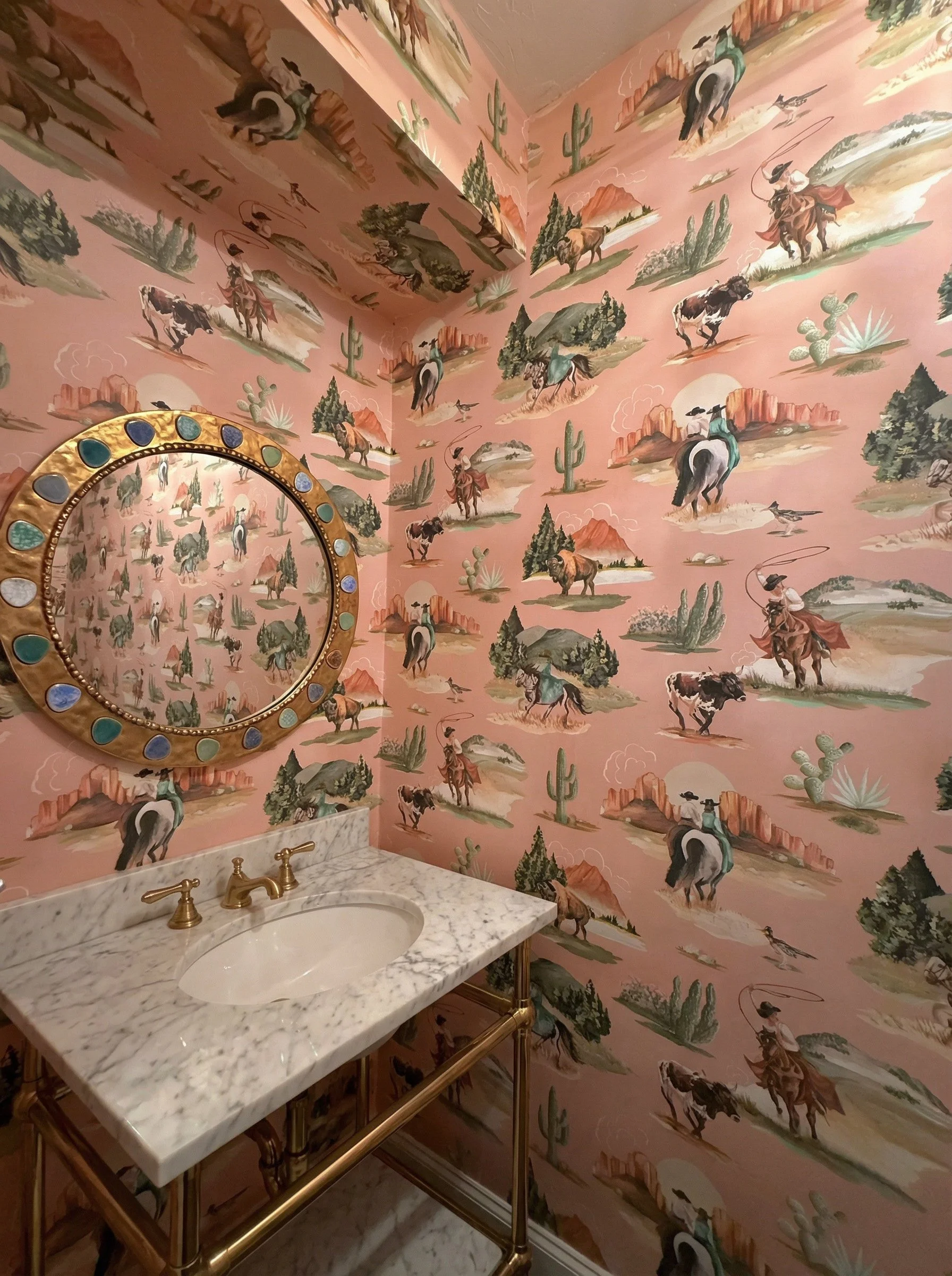 A bathroom with pink desert-themed wallpaper featuring cowboys, horses, cacti, and mountains, a marble countertop with a built-in sink, and a decorative mirror with a gold frame decorated with colorful stones.