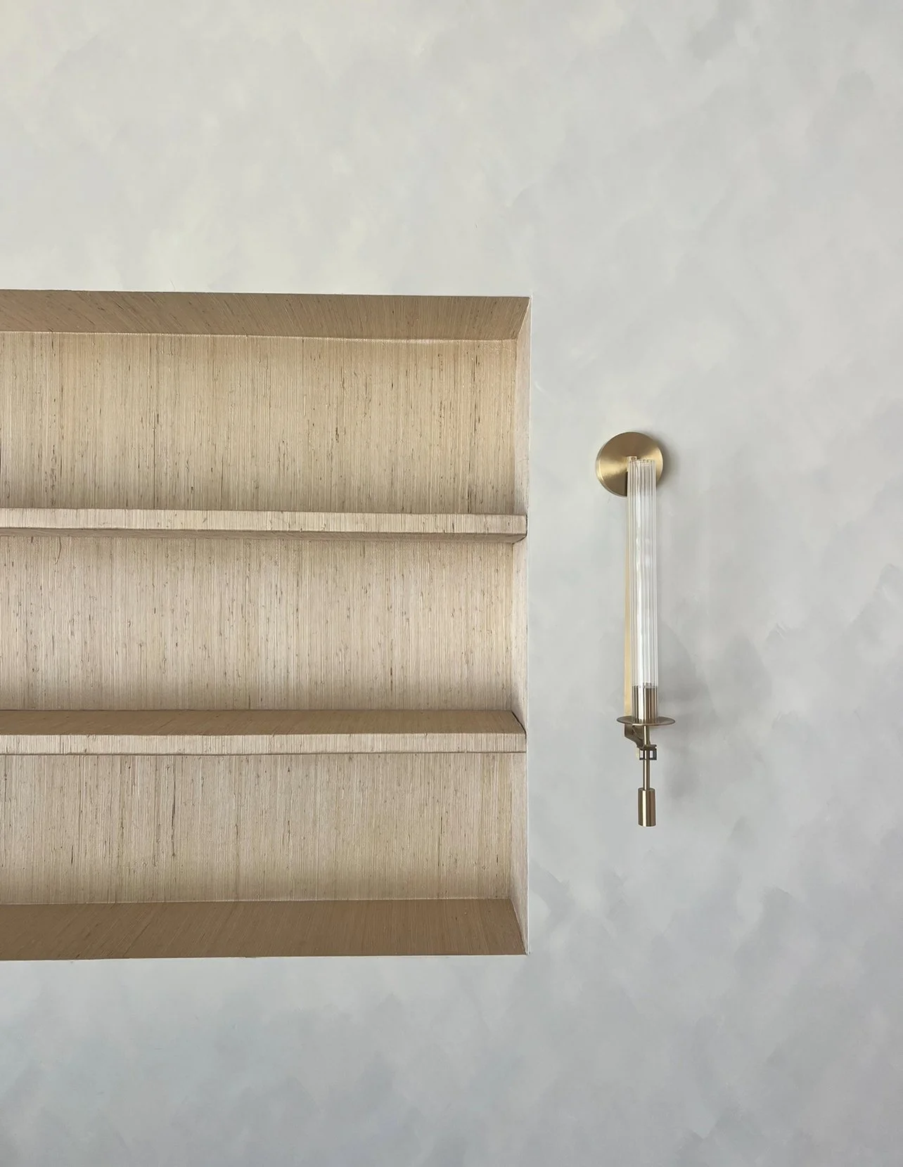 A minimalist wooden bookshelf with open shelves mounted on a light-colored wall, with a modern, vertical wall sconce to the right of the shelf.