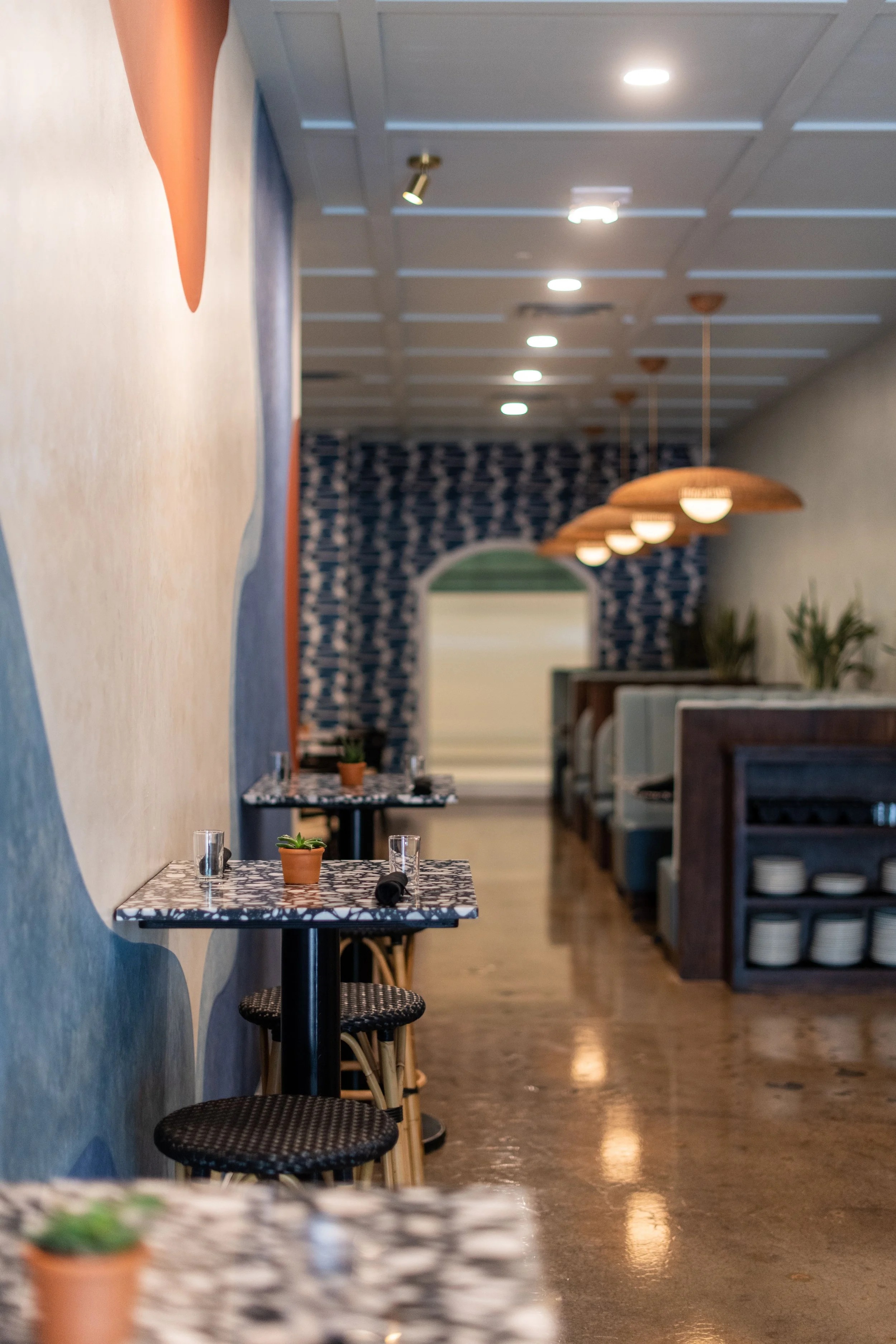Interior of a modern restaurant with tables, chairs, potted plants, and pendant lamps.