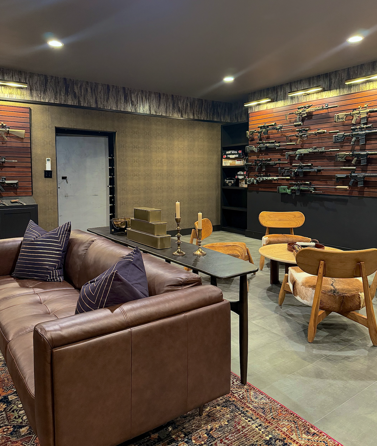 Interior of a room with a brown leather couch, wooden chairs, and a wall display of various guns. There are decorative candles on a black table, and shelves with boxes on the back wall.