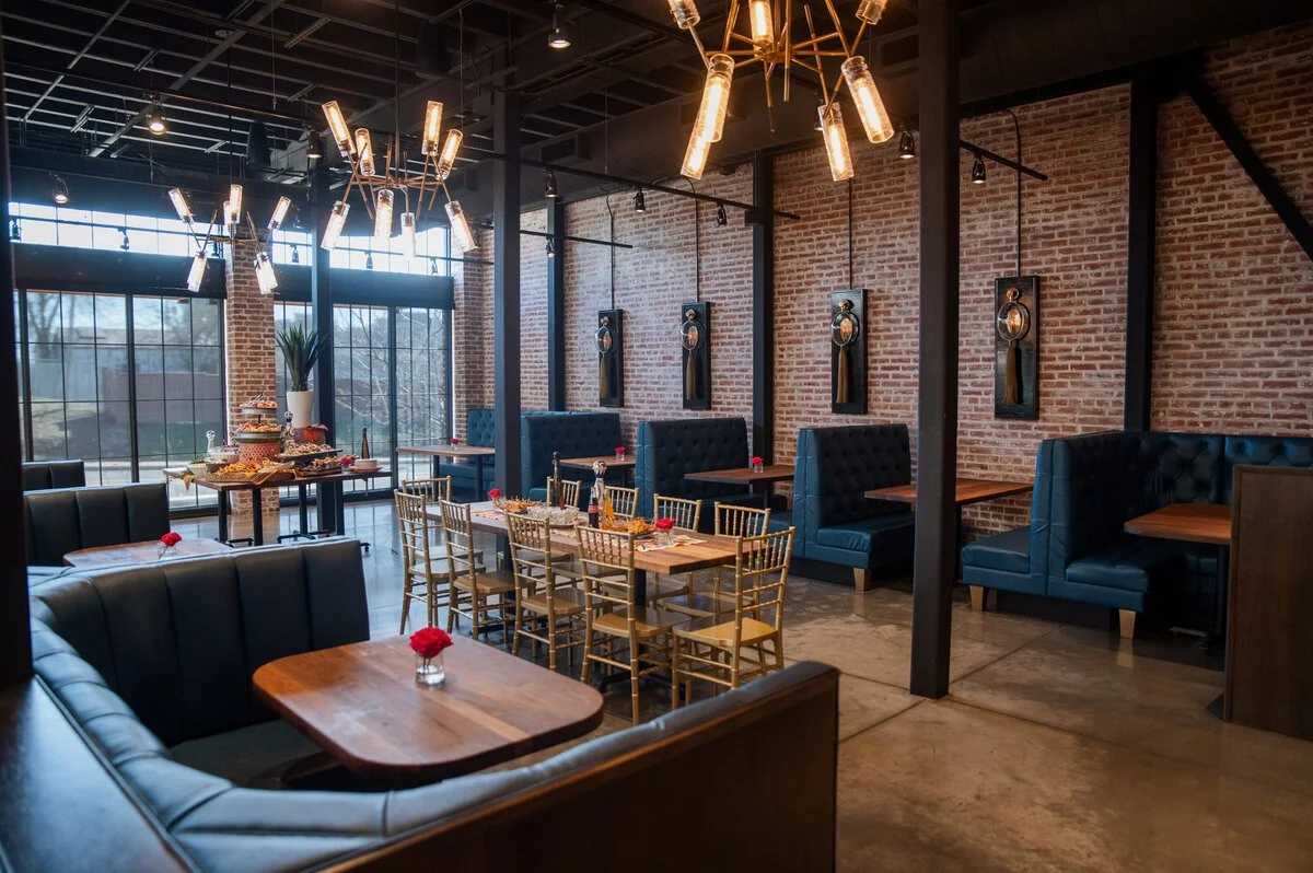 Interior of a modern restaurant with exposed brick walls, large windows, elegant black and gold lighting fixtures, blue cushioned booth seating, gold Chiavari chairs, a table with a dessert display, and small red flowers on each table.