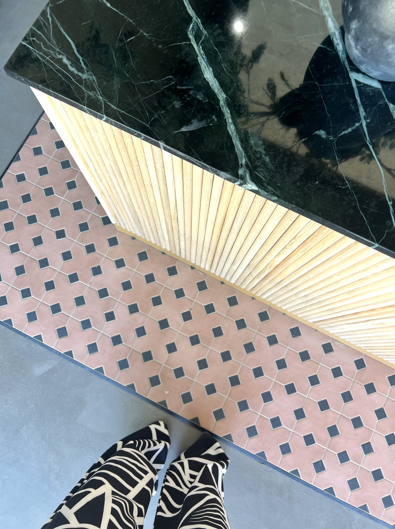 Photograph of a person standing on the sidewalk, showing their black and white patterned pants, next to a tiled floor, a wooden slatted cabinet, and a black marble countertop with white veining.