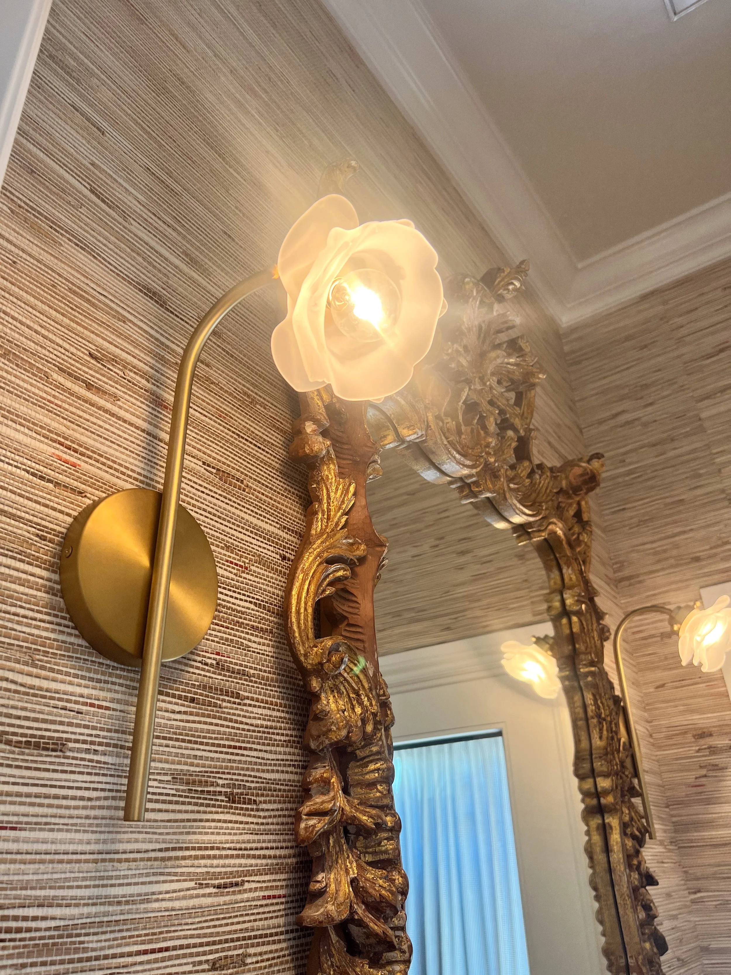 A wall-mounted, decorative brass light fixture with a floral-shaped glass lampshade, mounted beside an ornate, gilded mirror and a textured, tan-colored wallpaper.