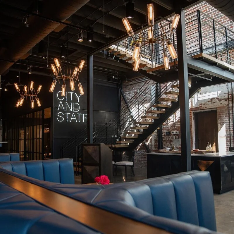 Interior of a modern restaurant or cafe with blue booth seating, exposed brick walls, a staircase with black metal railing, and contemporary hanging light fixtures.