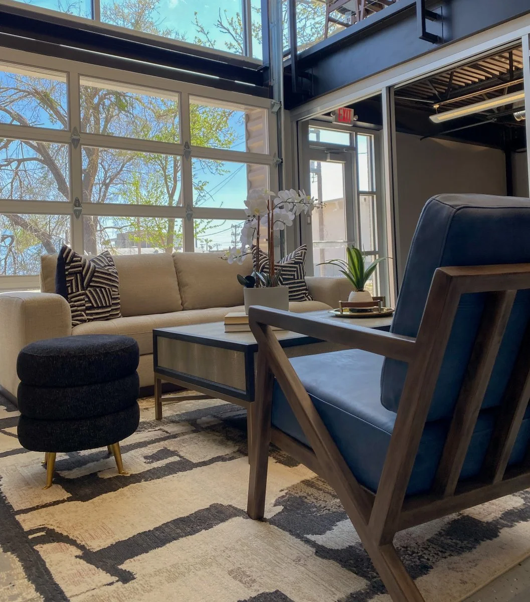 Modern lounge area with a beige sofa, black and white pillows, a coffee table with books and plants, a blue armchair, and a black round ottoman with gold legs, in front of large windows showing trees outside.