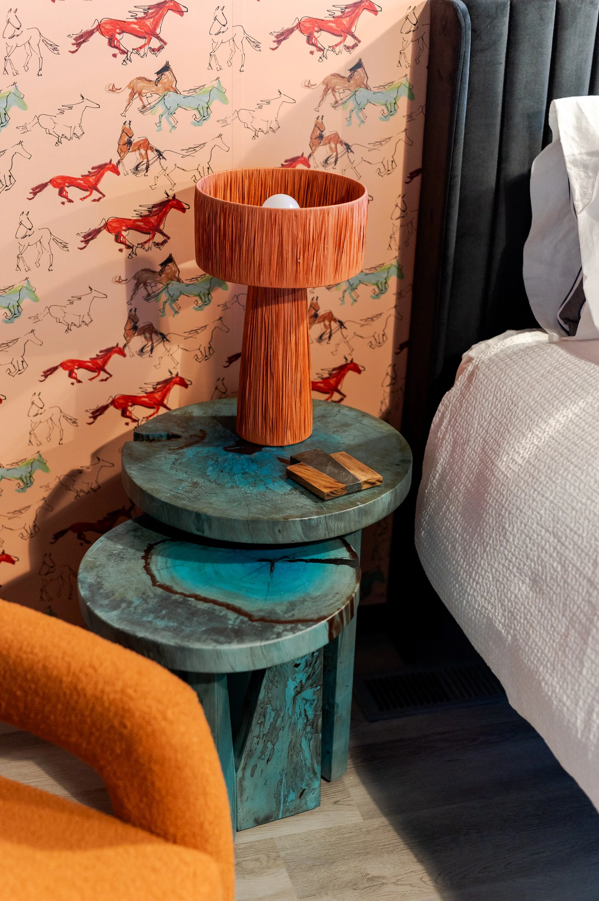 A bedroom nightstand with a wooden lamp, two round tables with blue-green marbled surfaces, and a small wooden tray. The wall behind features colorful horse sketches.