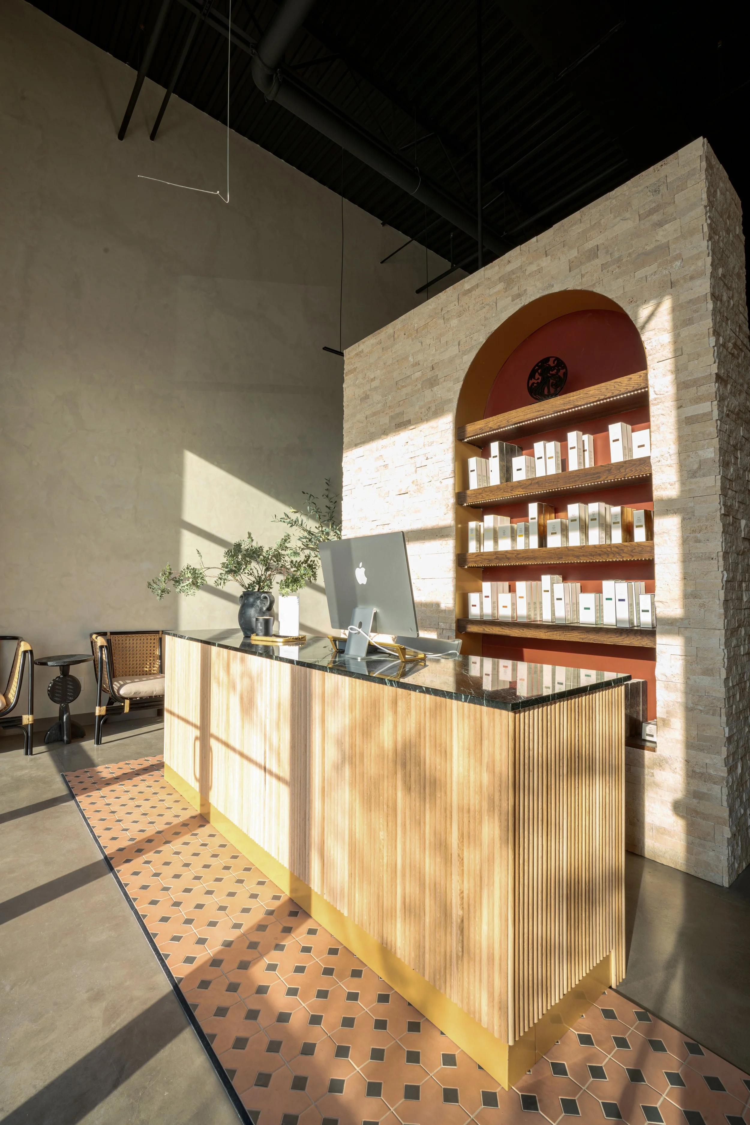 Modern reception desk with an Apple computer, decorative plants, and a brick wall with built-in shelves behind it, illuminated by sunlight.