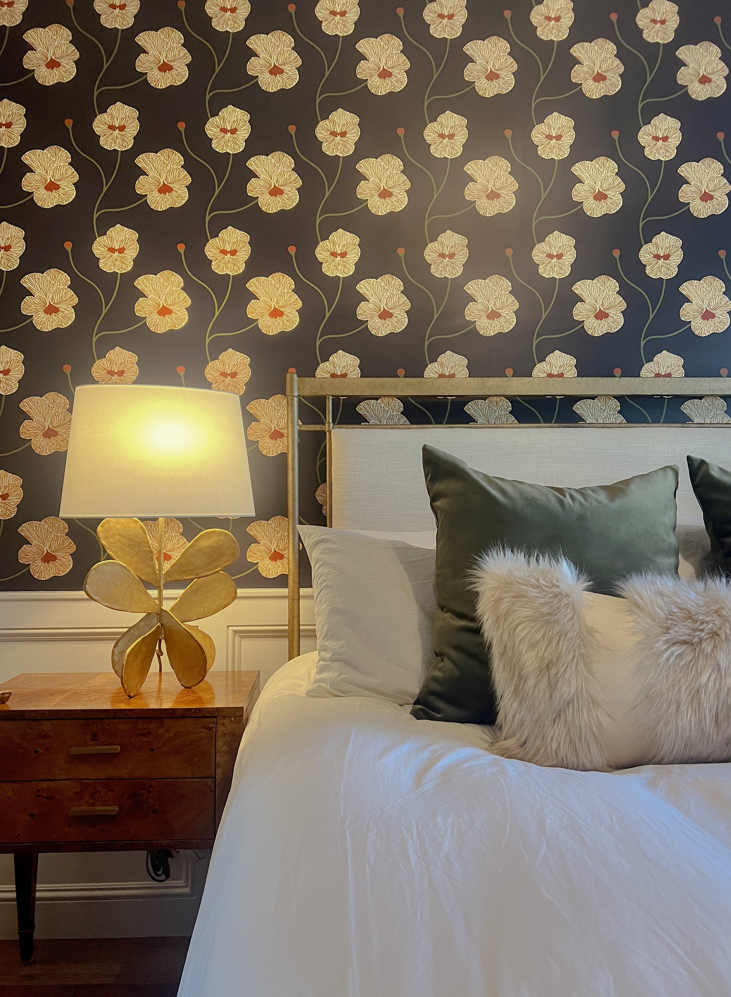 A bedroom with dark floral wallpaper, a beige headboard, green and white pillows with furry accents, a wooden nightstand, and a lamp with a flower-shaped base.
