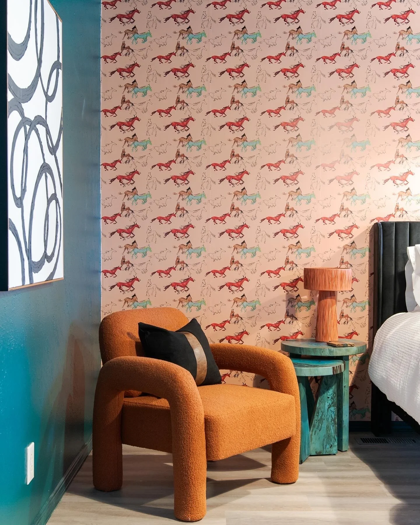 A cozy corner of a bedroom with a plush orange armchair, black pillow, teal side table with a wooden lamp, and a patterned wallpaper featuring colorful running horses.