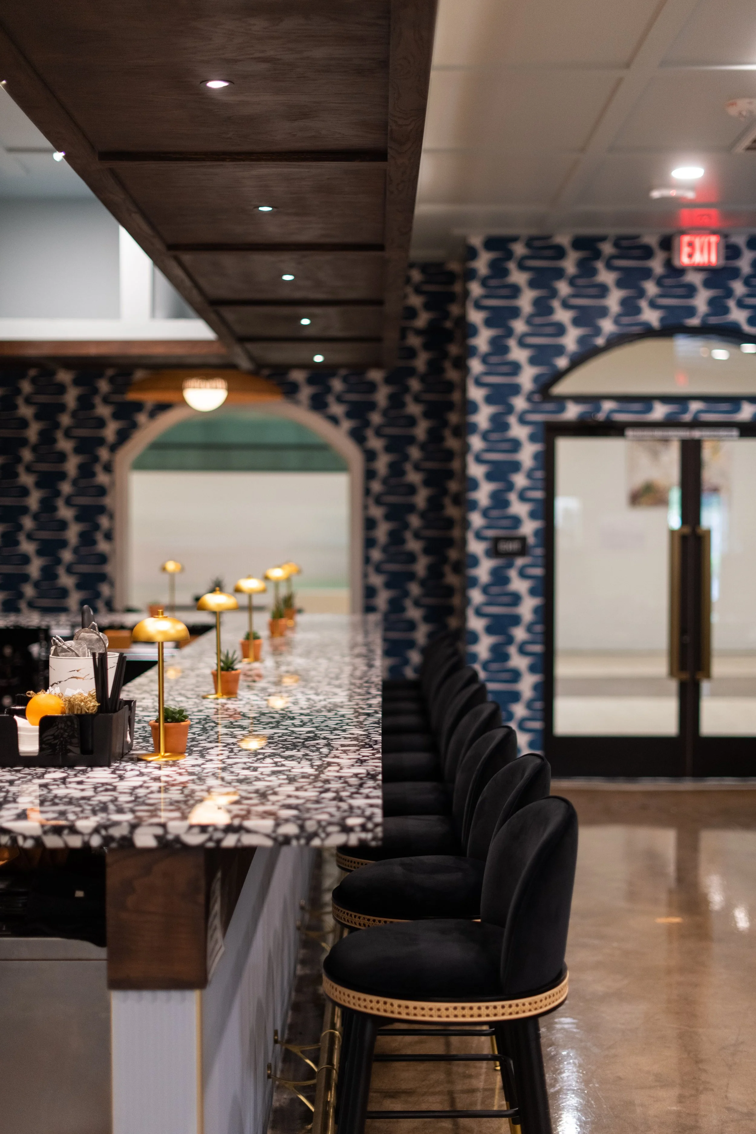 Interior of a bar or restaurant with a marble countertop, black bar stools, and decorative wallpaper with a blue and white pattern.