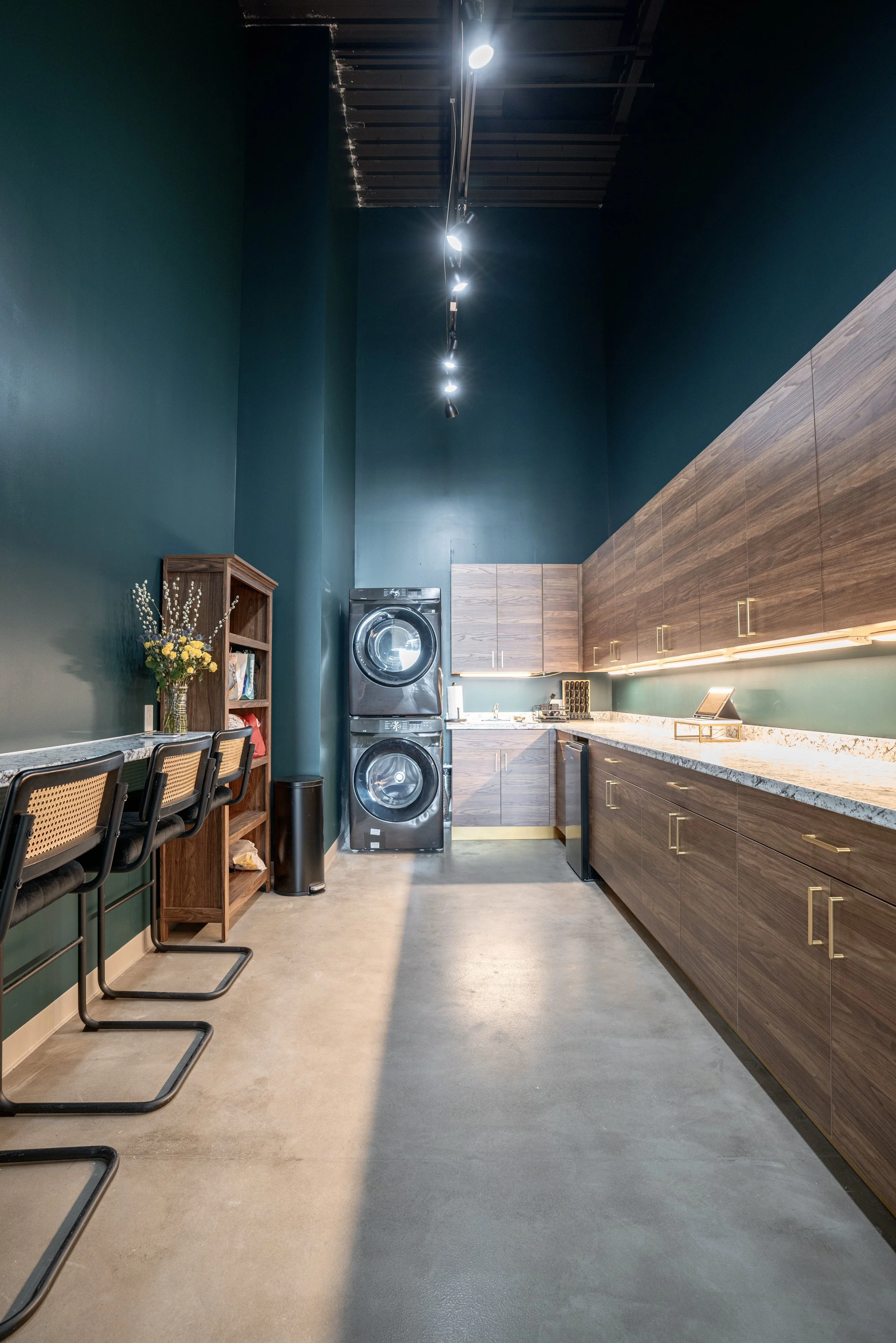 Modern laundry room with teal walls, wooden cabinets, a stacked washer and dryer, a small white sink, and a counter with under-cabinet lighting. There are three black chairs with cane backrests at a counter on the left, and a wooden shelf with flower
