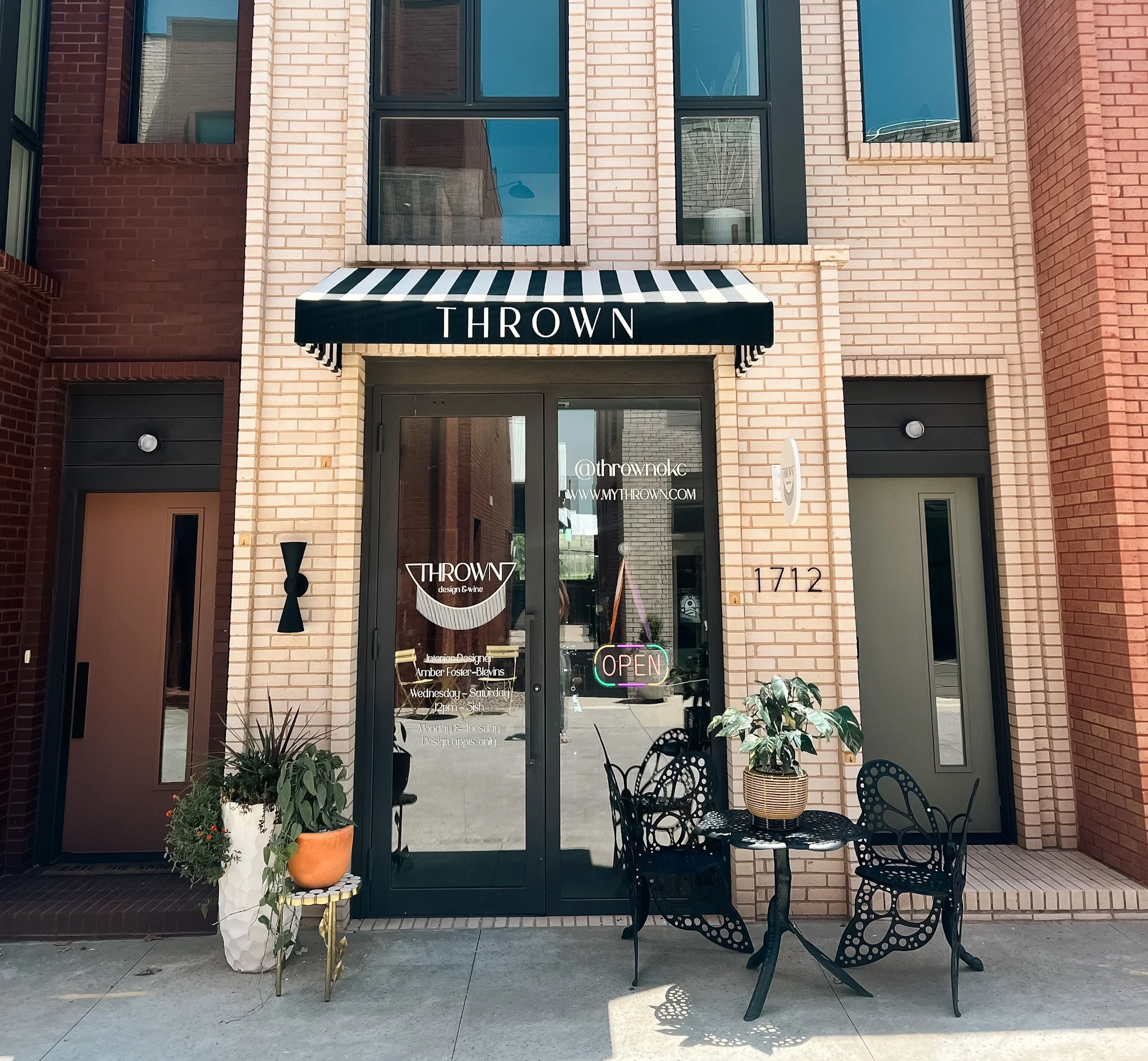 Exterior view of a modern brick building with a shop named 'THROWN', featuring glass door, black and white striped awning, and outdoor seating with black chairs and a small table, decorated with potted plants.