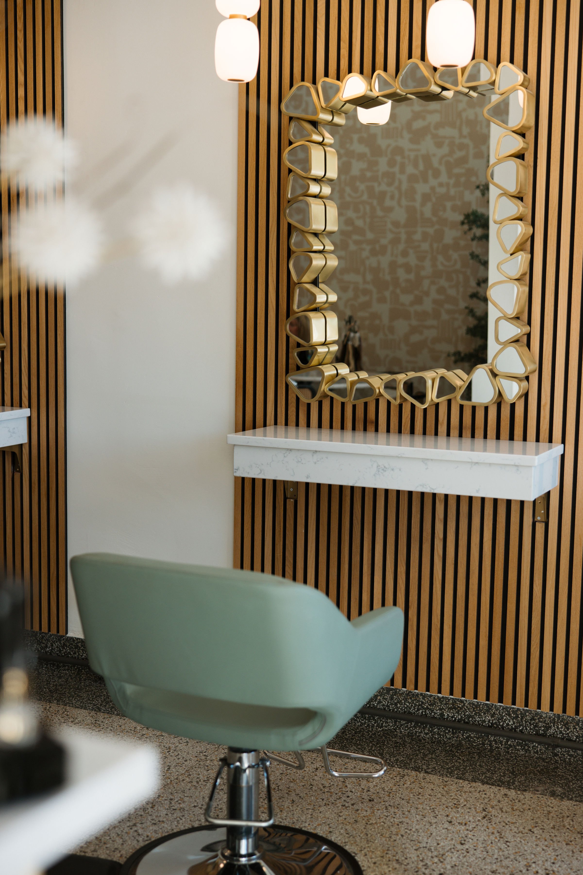 A modern salon interior featuring a large decorative mirror with a unique frame design, a white marble countertop, a green salon chair, warm lighting, wooden slat wall panels, and patterned wallpaper.