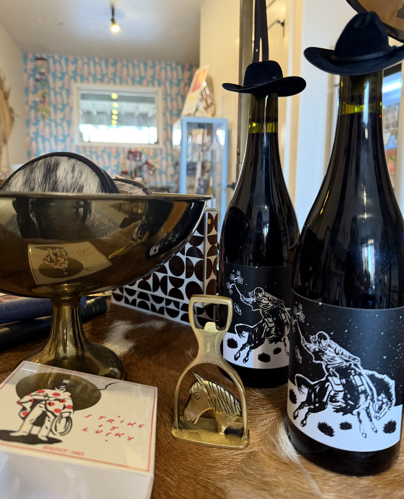 Two bottles of wine with cowboy hat labels, a gold-colored horse-shaped cork opener, and a small decorative bowl with a hat inside on a wooden table.