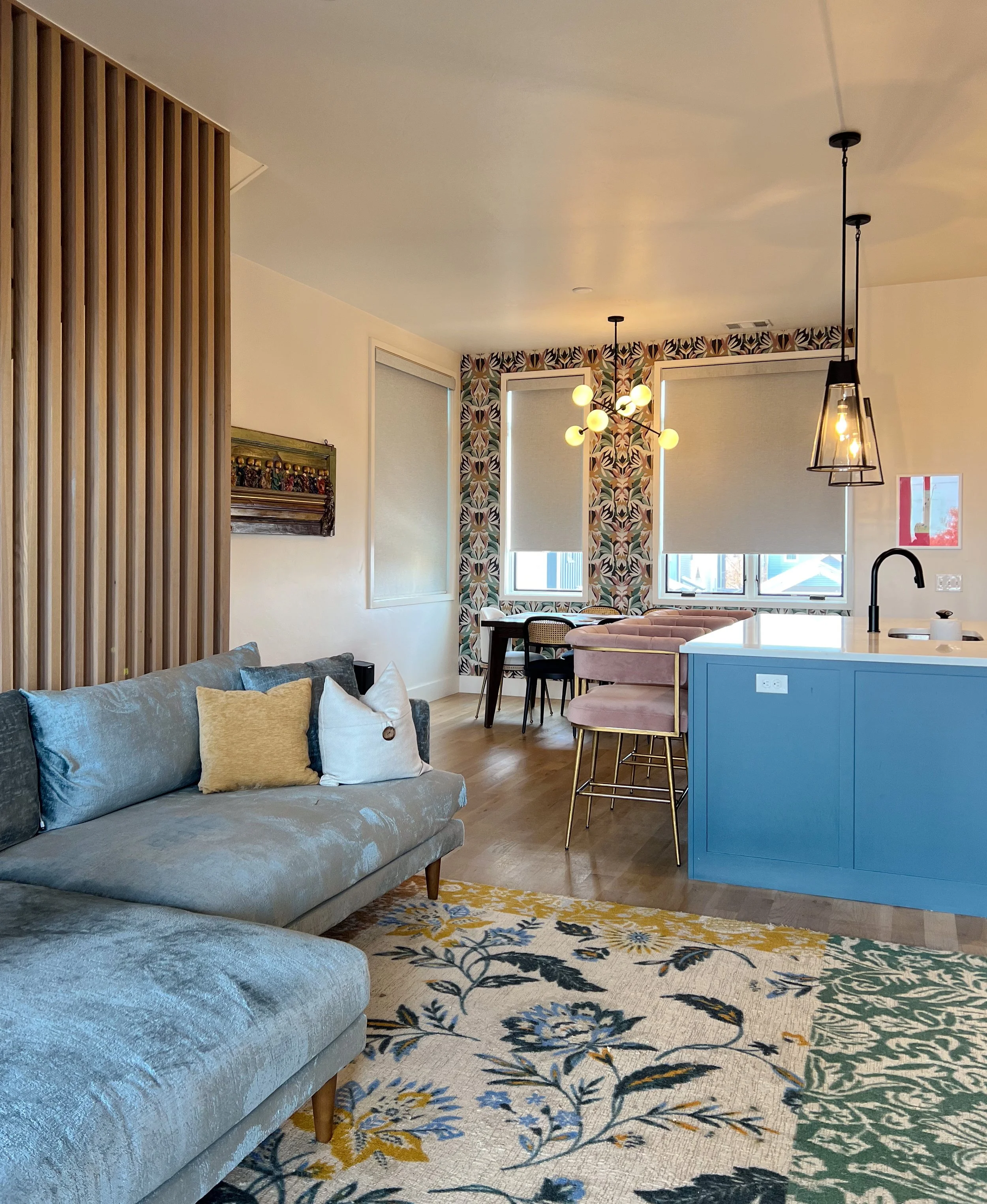 Living room with gray sofa, decorative pillows, wooden room divider, patterned area rug, and a dining area with pink chairs, a table, and windows with roller blinds; modern light fixtures hanging from ceiling.
