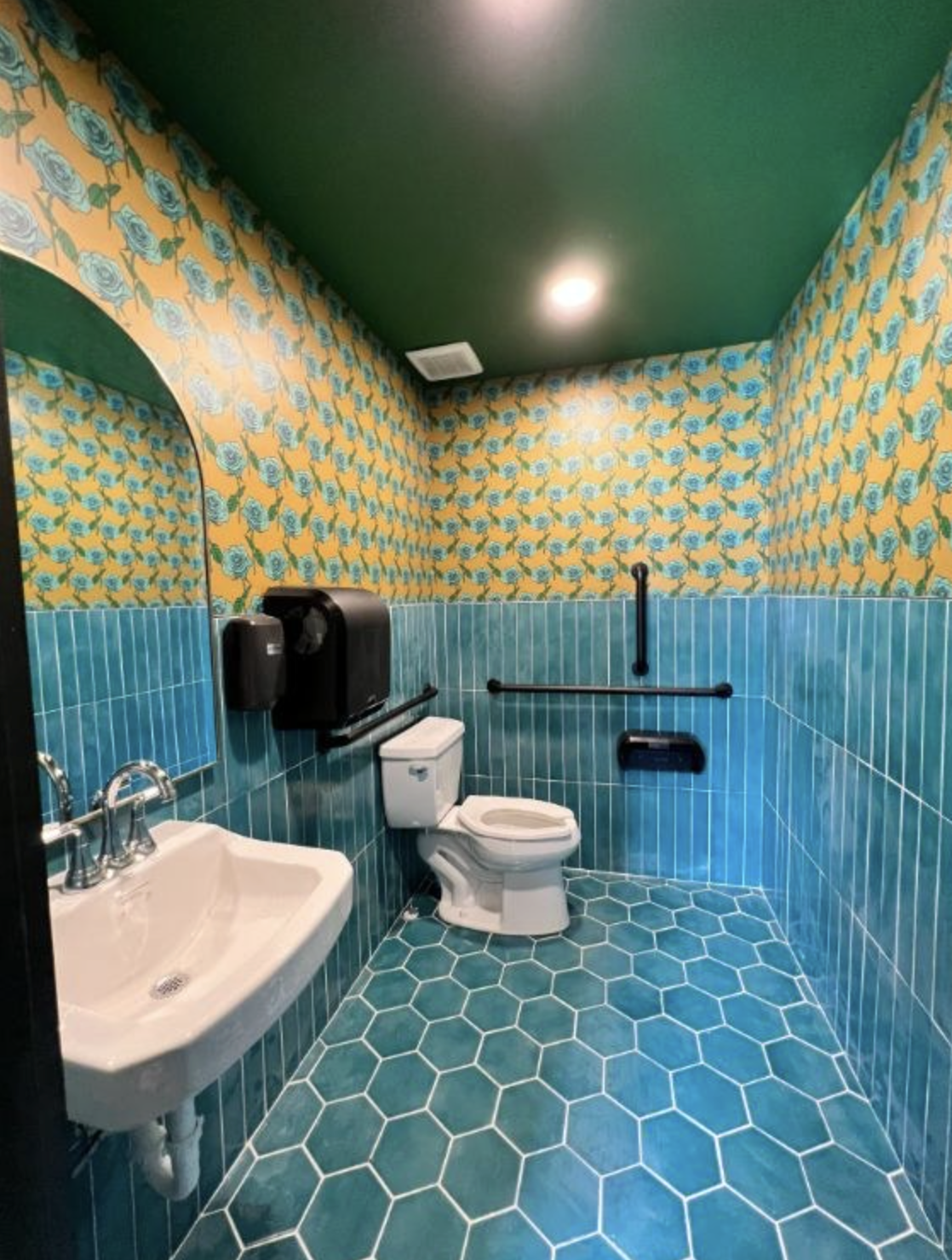 Bathroom with turquoise hexagon tile flooring, blue-green tiled walls, a white toilet, a white sink, a mirror, and sunflower wallpaper with blue roses on yellow background. Green ceiling with two ceiling lights.