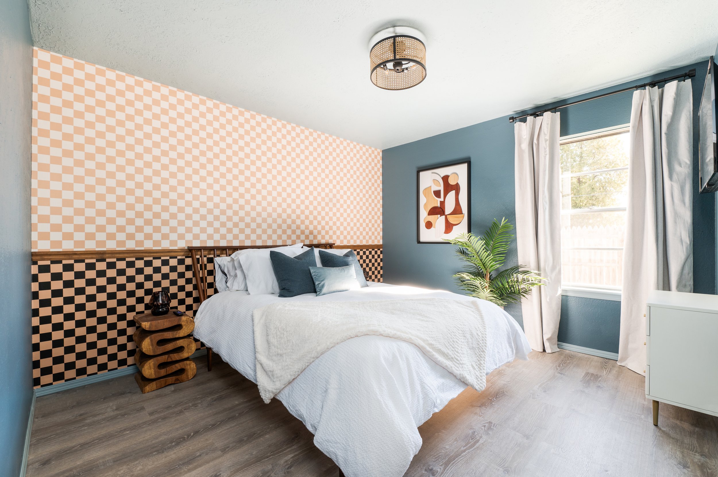 A bedroom with a white bed, dark blue pillows, a patterned accent wall, a framed abstract art piece, a window with white curtains, and a potted plant.