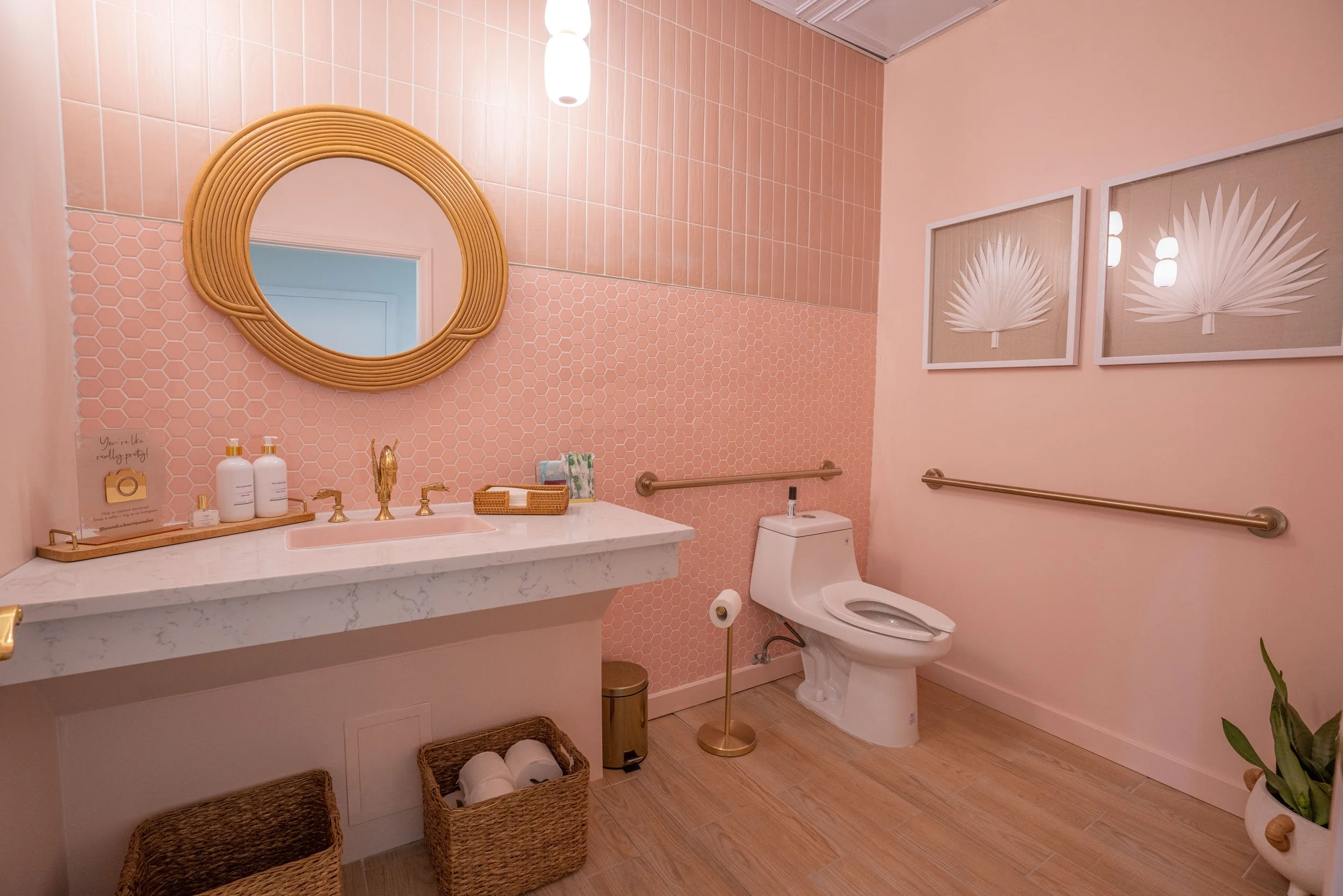 Pink-themed bathroom with a marble vanity, round mirror, gold fixtures, framed leaf artwork, and a white toilet.