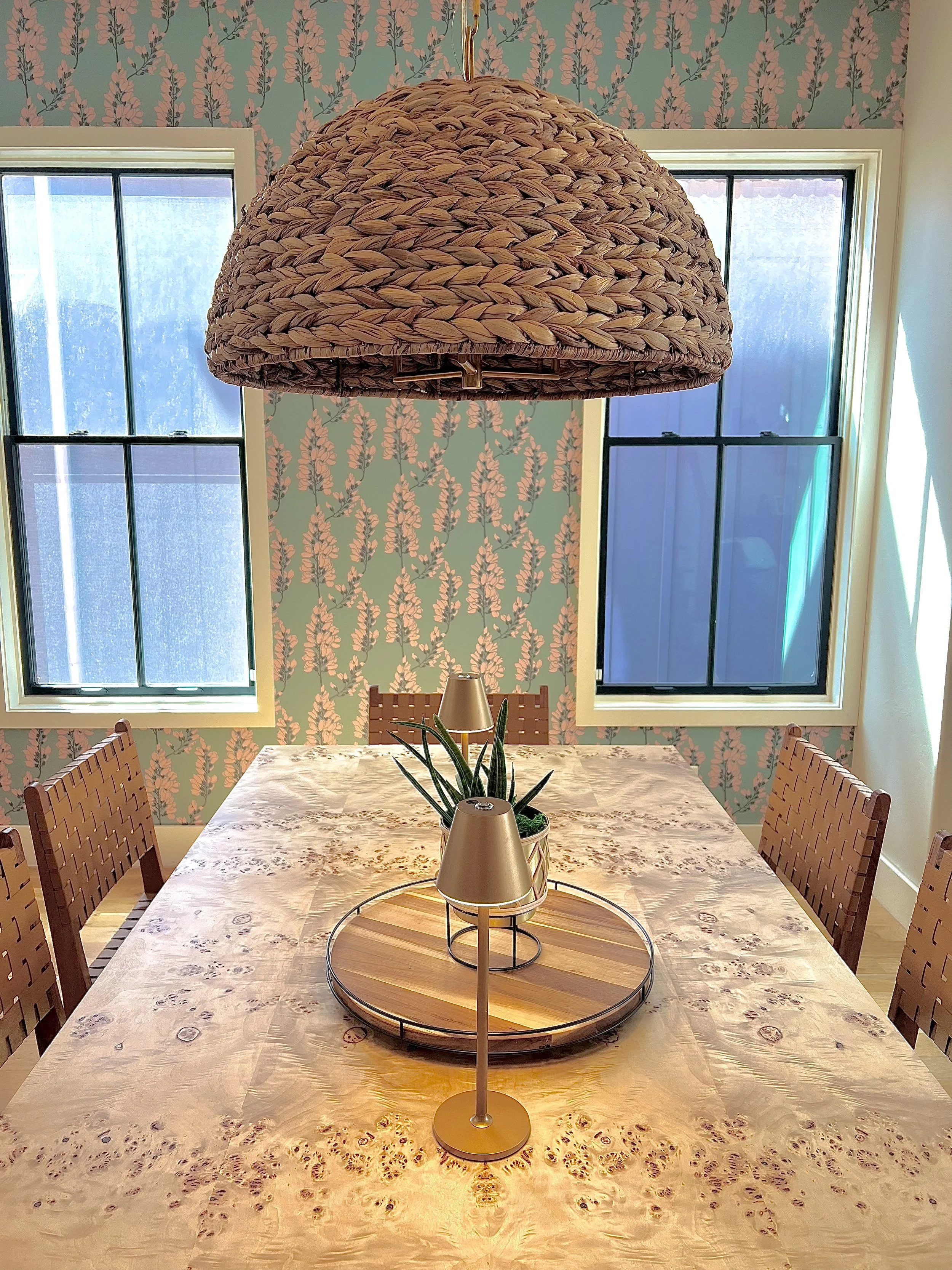 A dining room with a large woven pendant light hanging above a light-colored, polished wooden table with a burl wood pattern. The table has a tray with a potted plant and two small lamps. There are four wooden chairs with woven backs around the table
