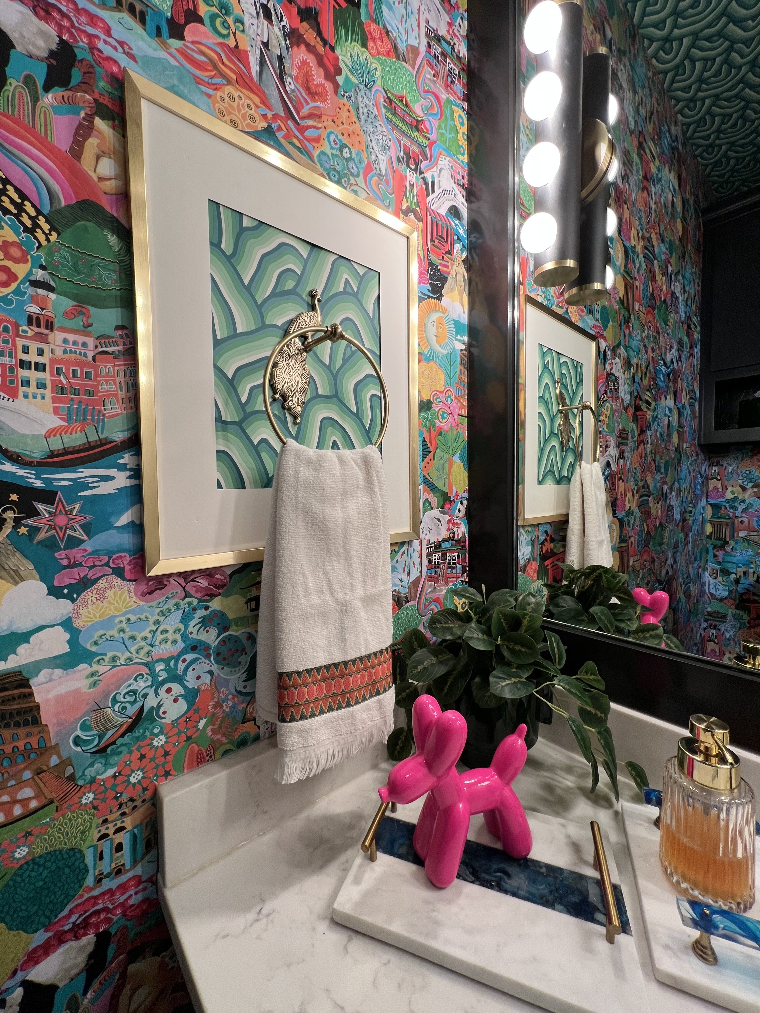 Bathroom with vibrant, colorful wallpaper featuring abstract and scenic designs, a large mirror, modern light fixture, artwork with a bird ornament, a pink balloon dog sculpture, a potted plant, and a marble countertop with a soap dispenser.