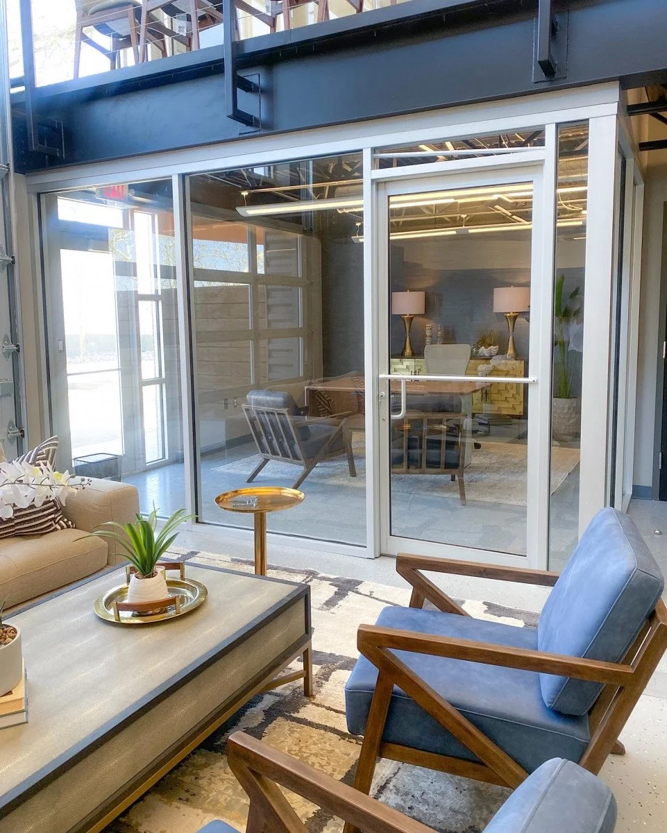 A modern indoor-outdoor workspace or lounge area with glass walls and furniture, including a beige sofa, blue armchairs, a coffee table with a plant, and a separate office space inside visible through the glass.