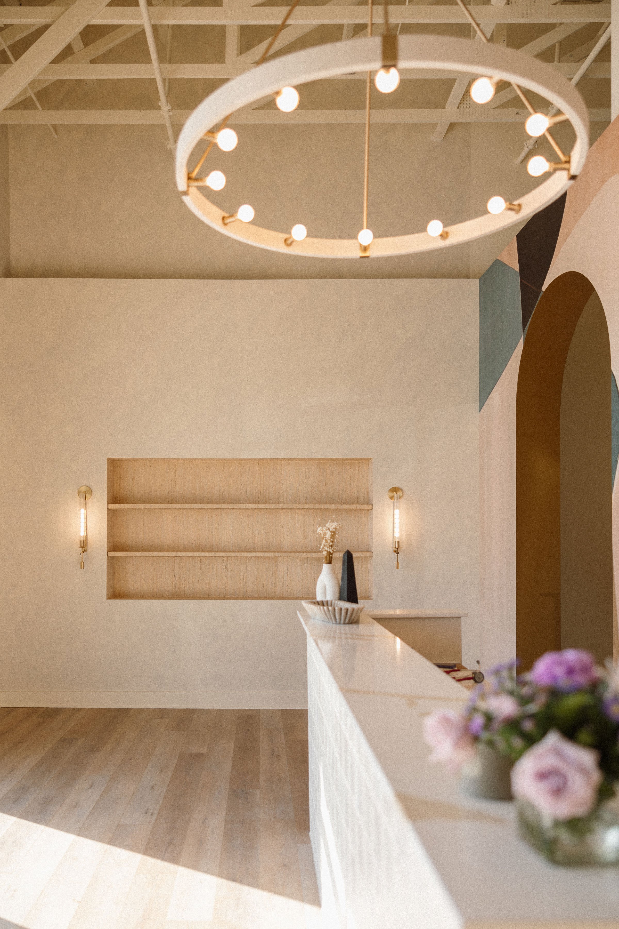 Minimalist interior with a circular chandelier, wooden floor, built-in shelves, and wall sconces, decorated with vases and flowers.