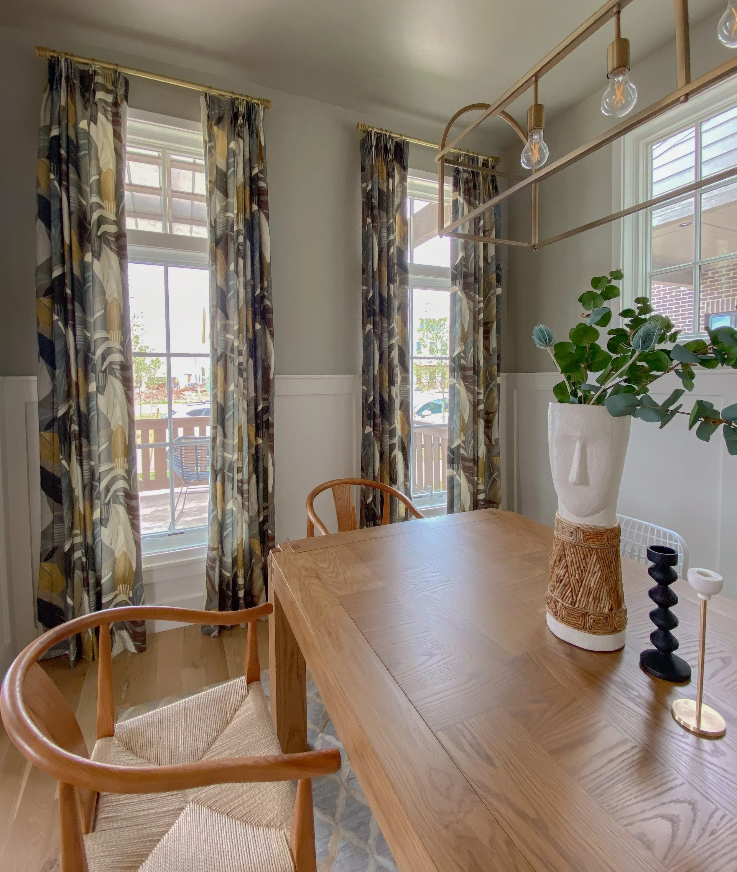 A dining room with a wooden table, chairs, patterned curtains, and a decorative vase with greenery.