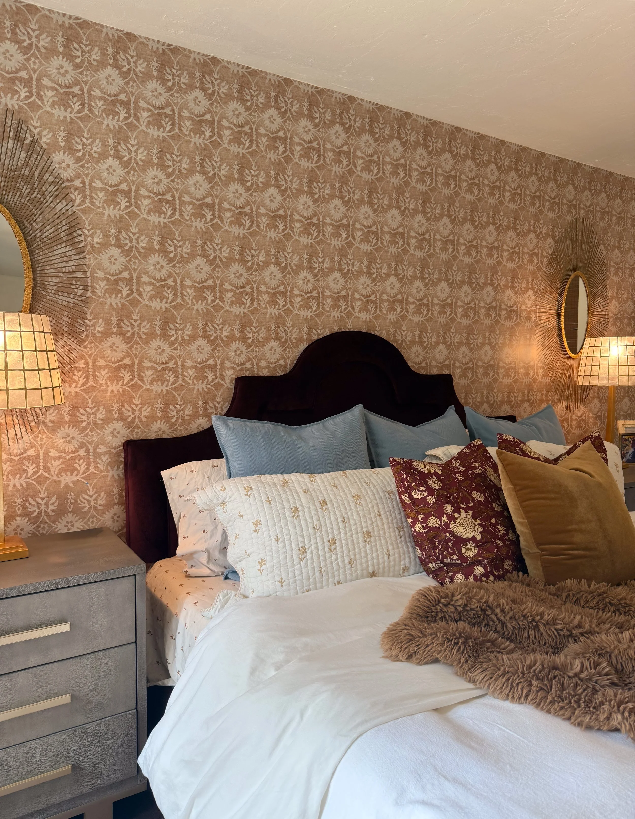 A neatly made bed with multiple pillows against a dark headboard, a nightstand with a lamp, and warm, decorative wallpaper with oval mirrors and lighting.