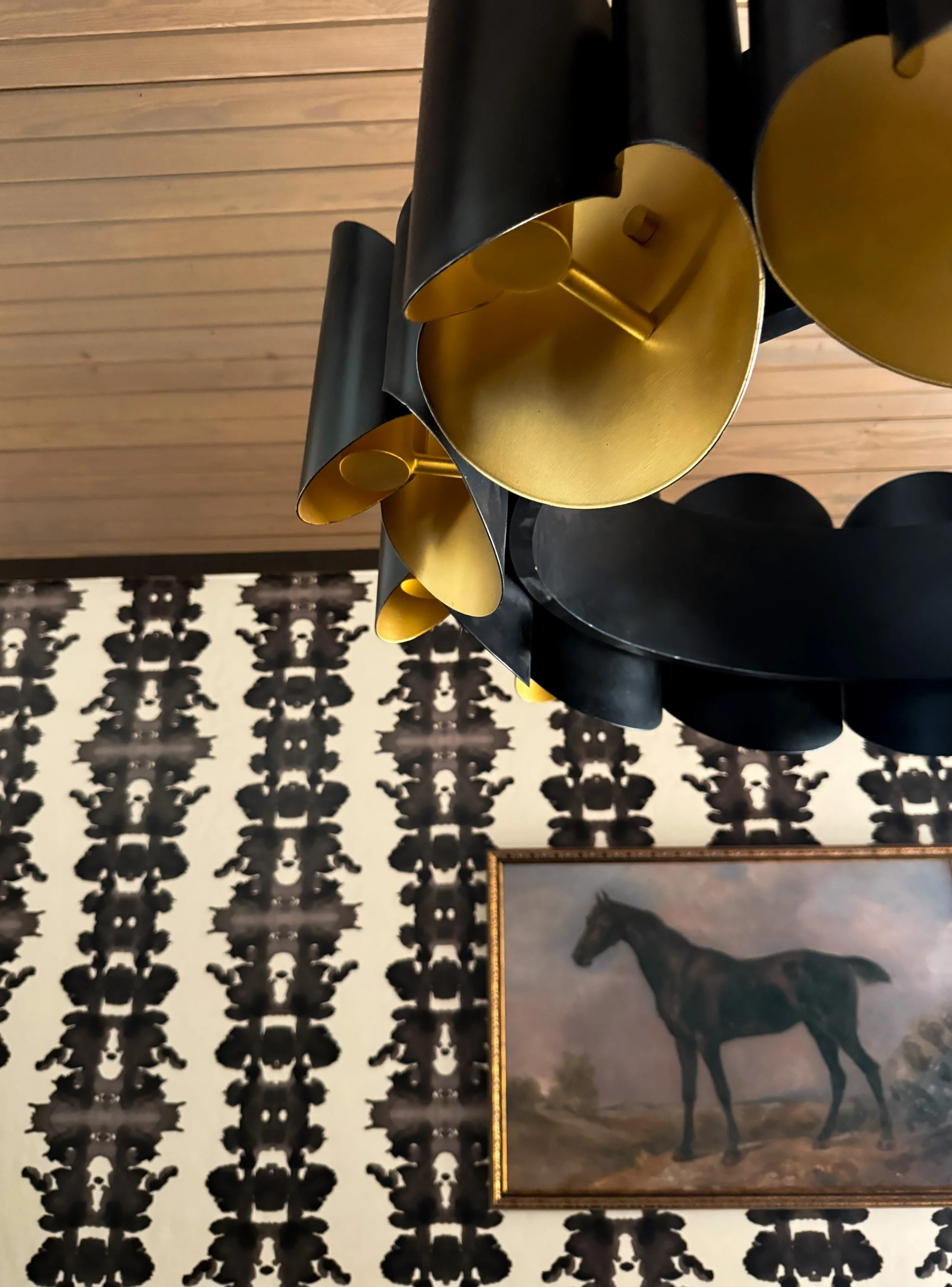 A modern black and gold chandelier hanging from a ceiling, with a framed painting of a horse on a patterned black and white wall in the background.