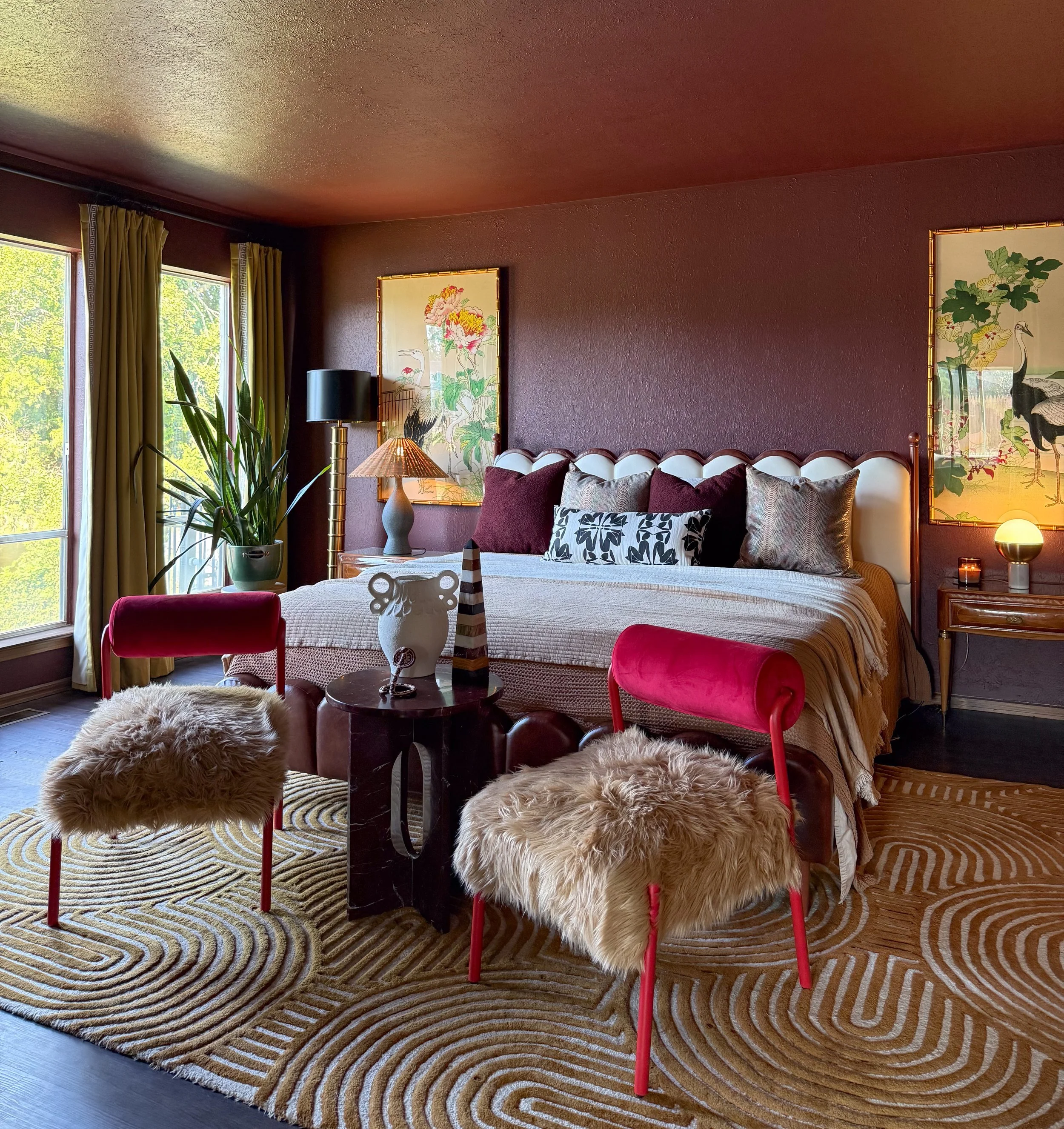 A bedroom with a large bed with multiple pillows, floral artwork on the walls, two chairs with faux fur cushions, a small round table with decorative vases, a tall plant near a window, and warm lighting.