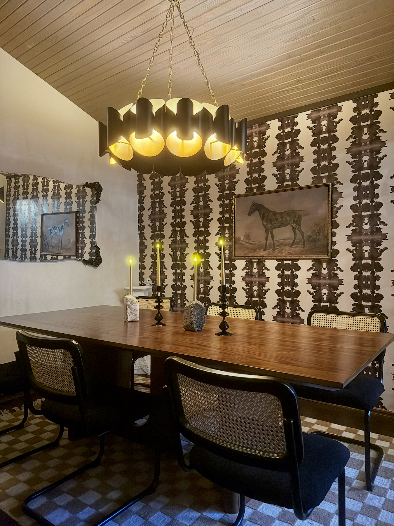 A dining room with a wooden table, six black chairs with cane backs, three lit candlesticks, and artwork of a horse on the patterned wallpapered wall.