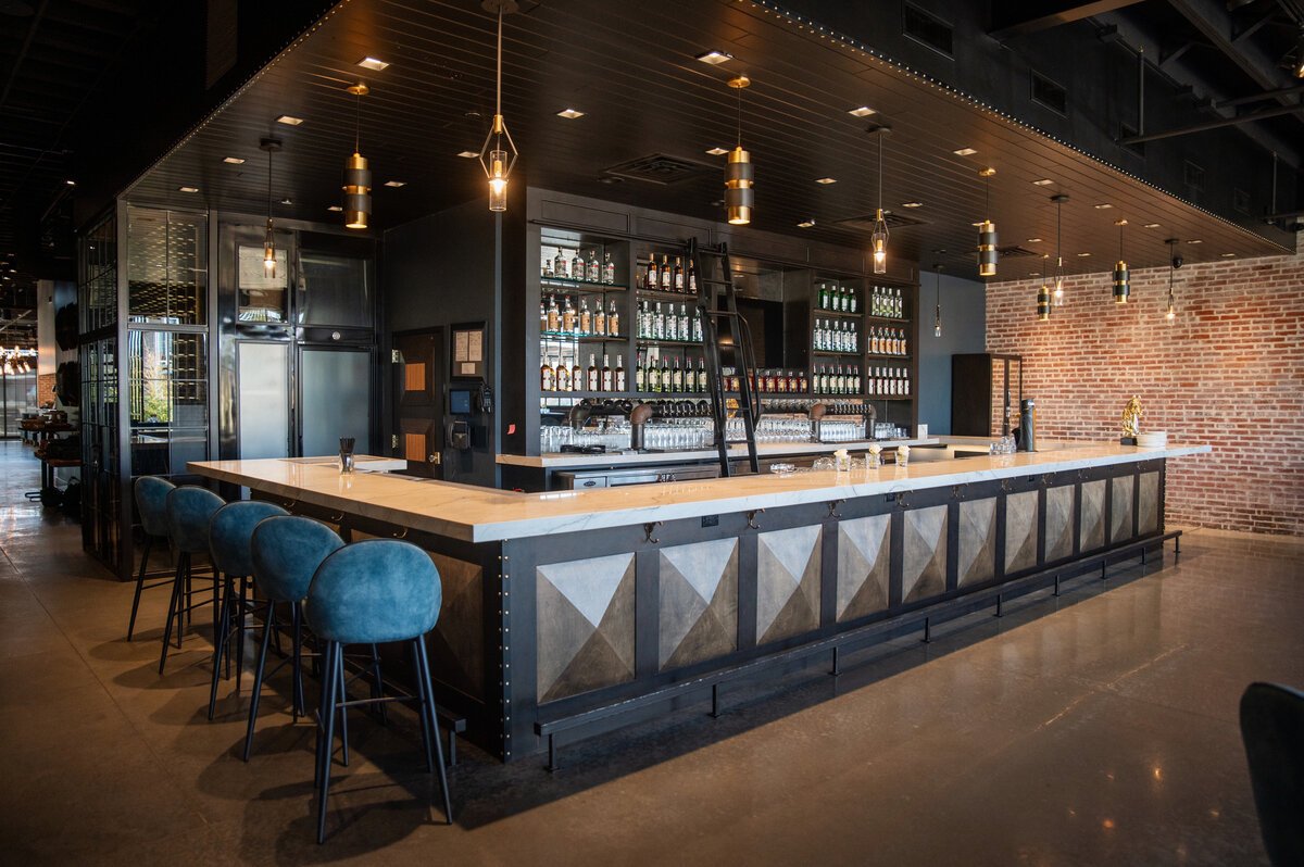 Modern bar with a white marble countertop, black and wood-paneled front, several blue bar stools, shelves with various bottles and glasses, hanging pendant lights, and a brick wall in the background.