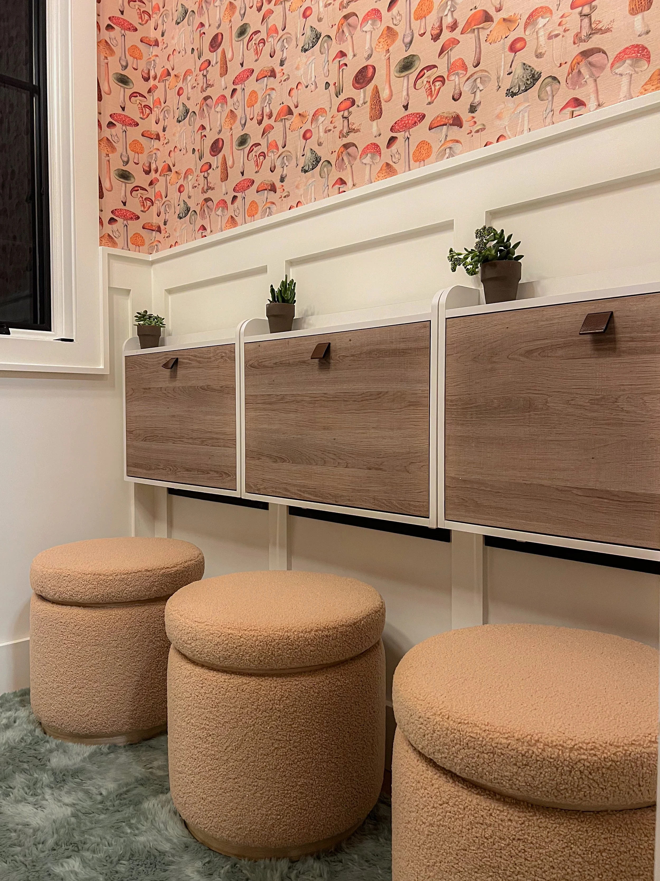 Interior of a cozy room with a wall decorated with pink mushroom-patterned wallpaper, a white shelf with small potted succulent plants, beige round upholstered stools, and a window with black frame.