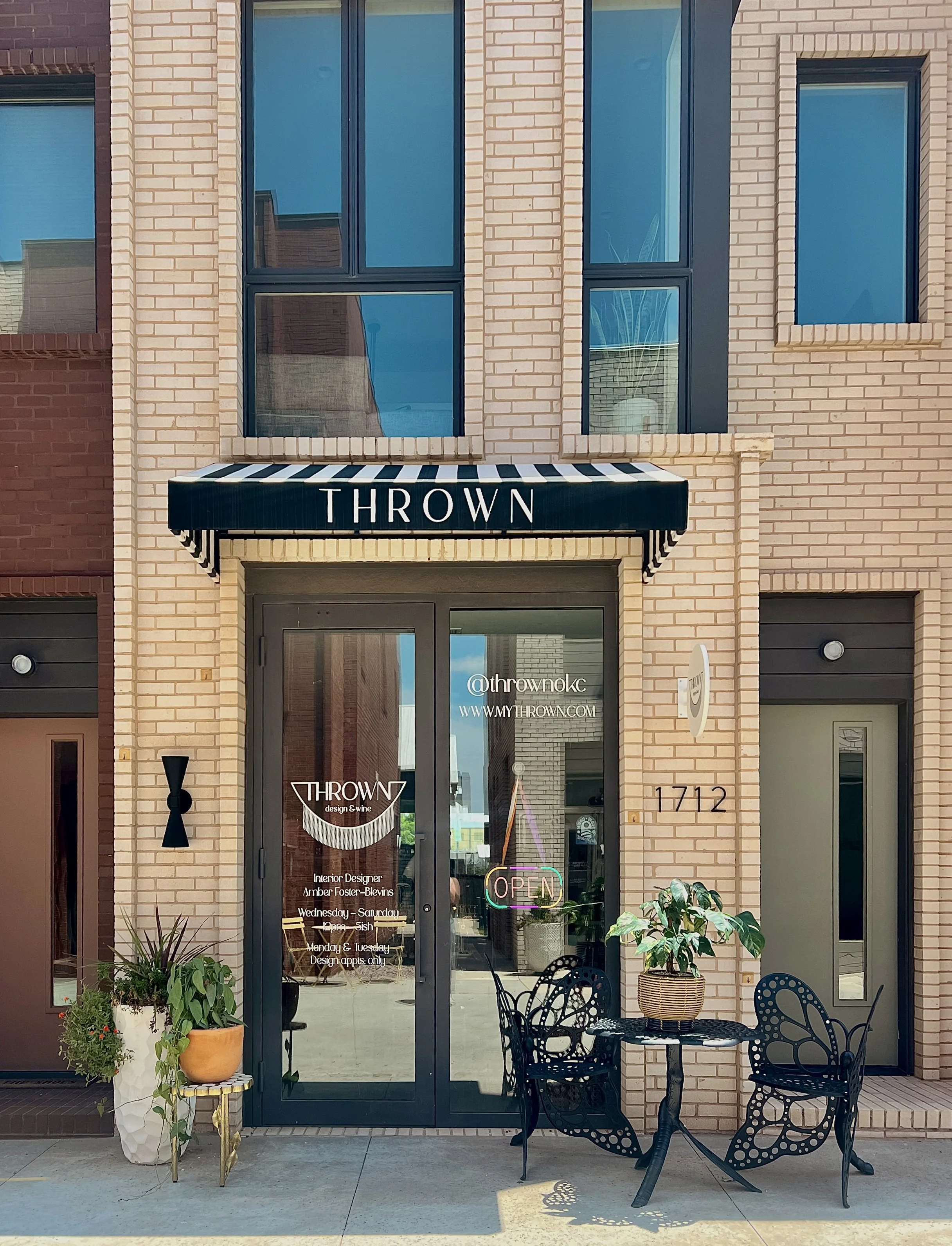 Exterior view of a modern brick building with a shop named 'THROWN', featuring glass door, black and white striped awning, and outdoor seating with black chairs and a small table, decorated with potted plants.