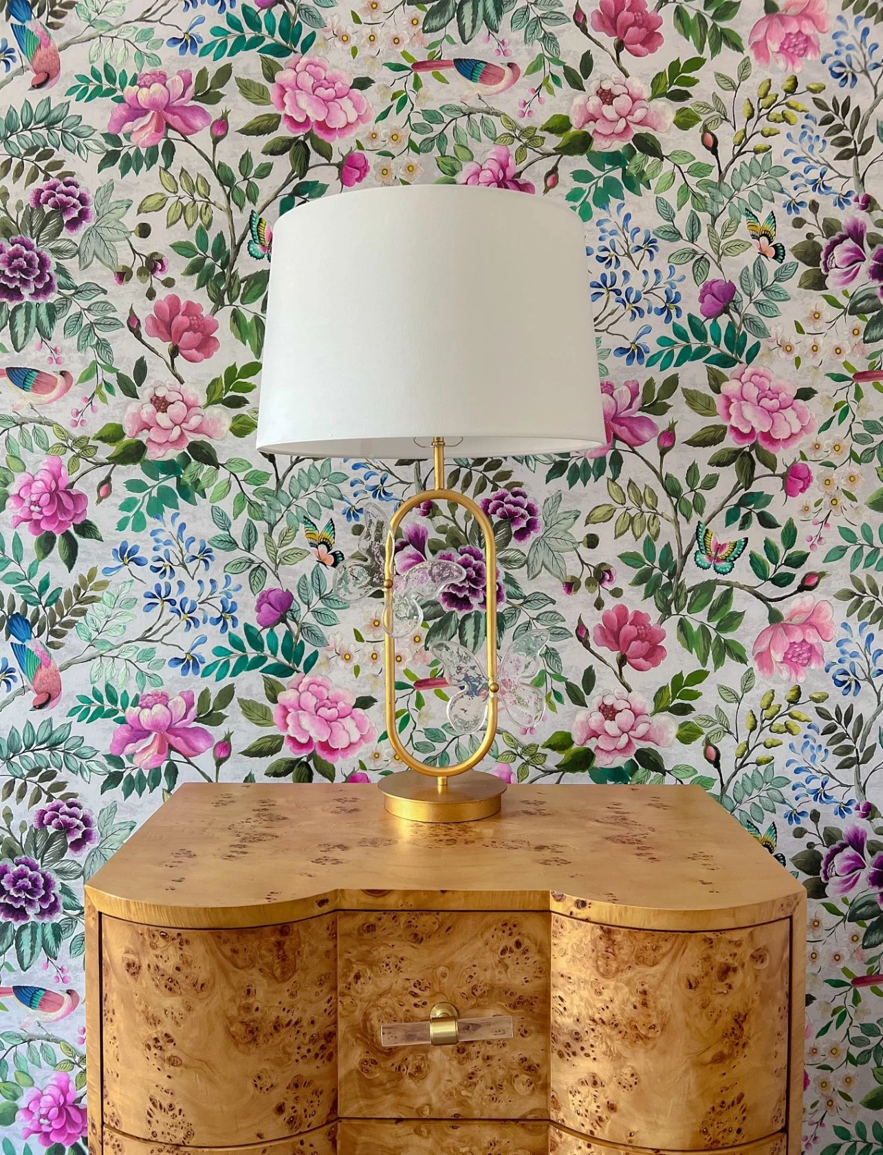 Table lamp with a gold base and a white shade, placed on a wooden table with a floral and butterfly wallpaper background.