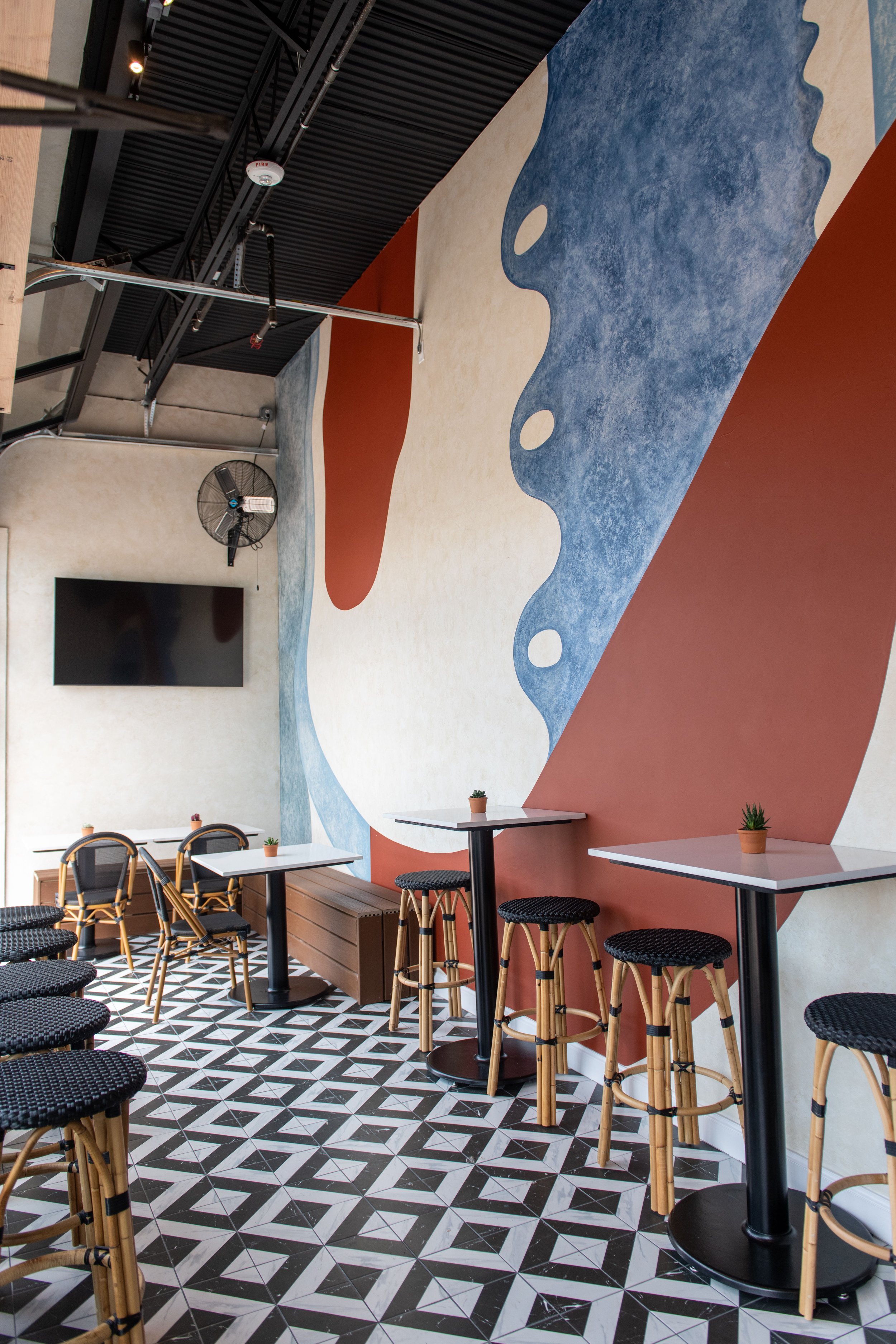 Interior of a modern cafe with high tables and bar stools, decorated with a large abstract mural on the wall and patterned tile floor.