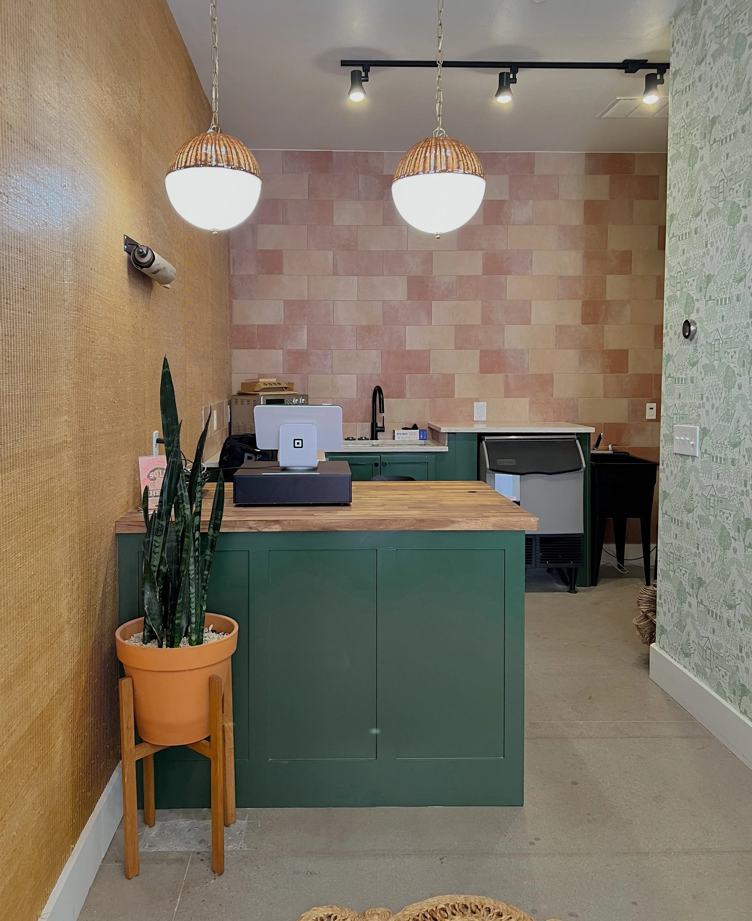 Interior of a kitchen with a green island, pink tile backsplash, and a black faucet. Decor includes two hanging light fixtures, a potted snake plant on a wooden stand, and a wall with colorful wallpaper.