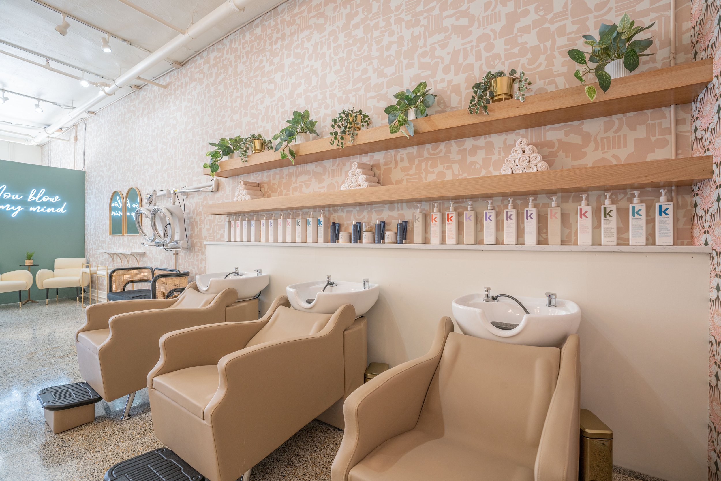 Interior of a modern hair salon with beige chairs at sinks, shelves with plants and hair products, and a waiting area with white chairs and a neon sign.