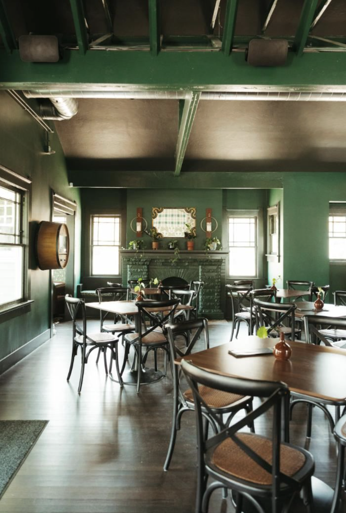 Interior of a cozy restaurant or cafe with green walls, wooden tables, black chairs with woven seats, and a fireplace decorated with plants and ornaments.