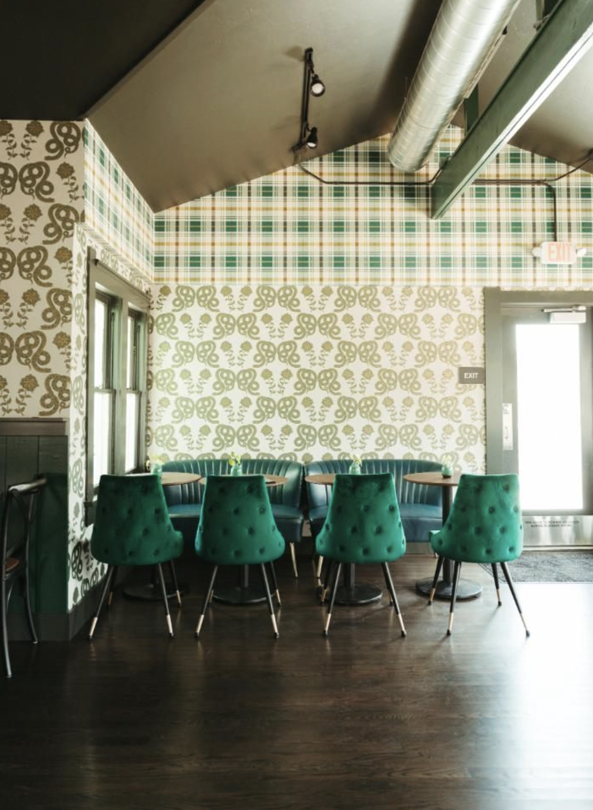 Interior of a cozy restaurant or cafe featuring a wall with floral-patterned wallpaper, green upholstered chairs, small round tables, and natural light coming through windows and a glass door.